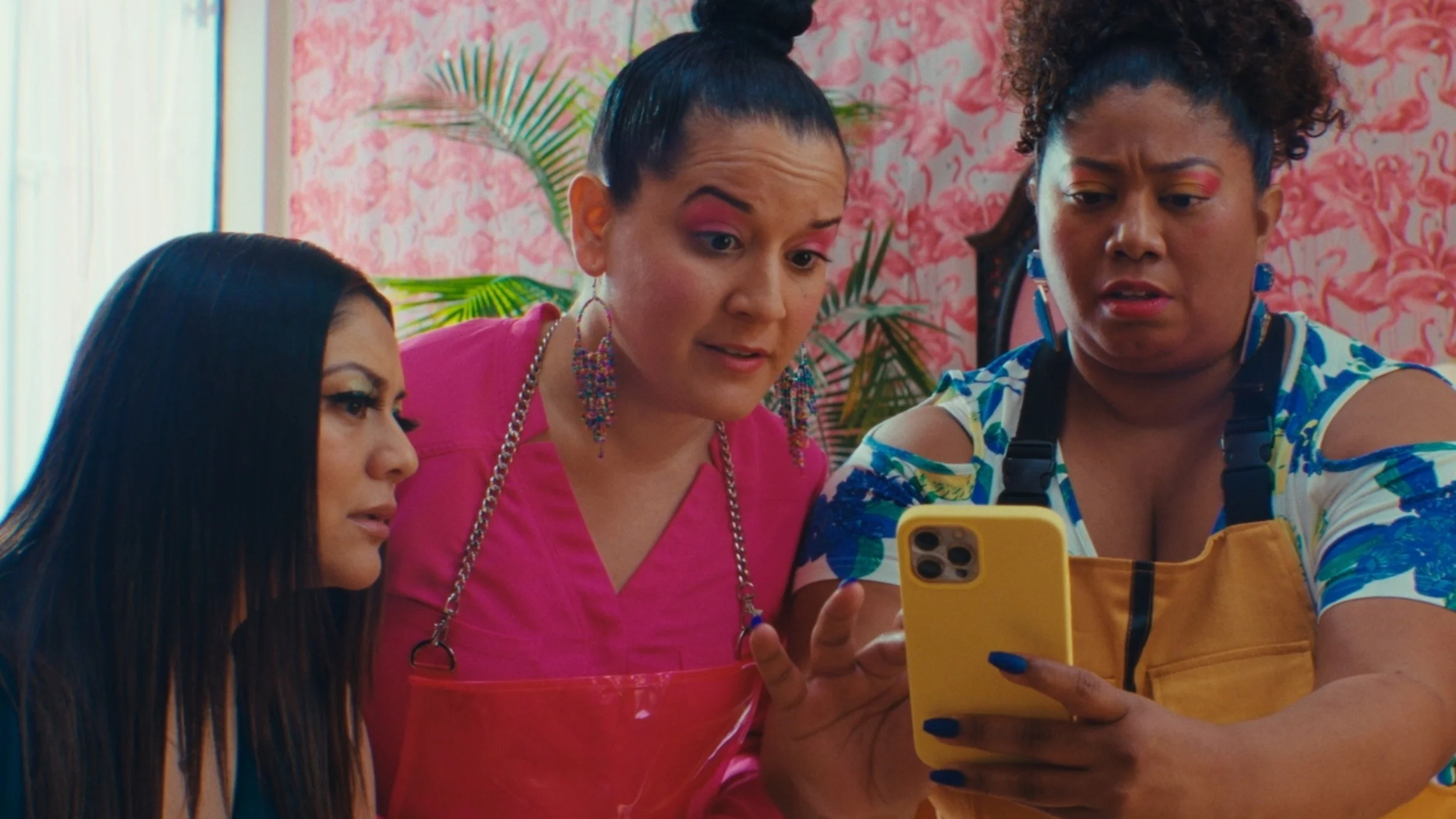 Three women with colorful makeup looking at a yellow smartphone, standing against a pink floral wallpaper background.