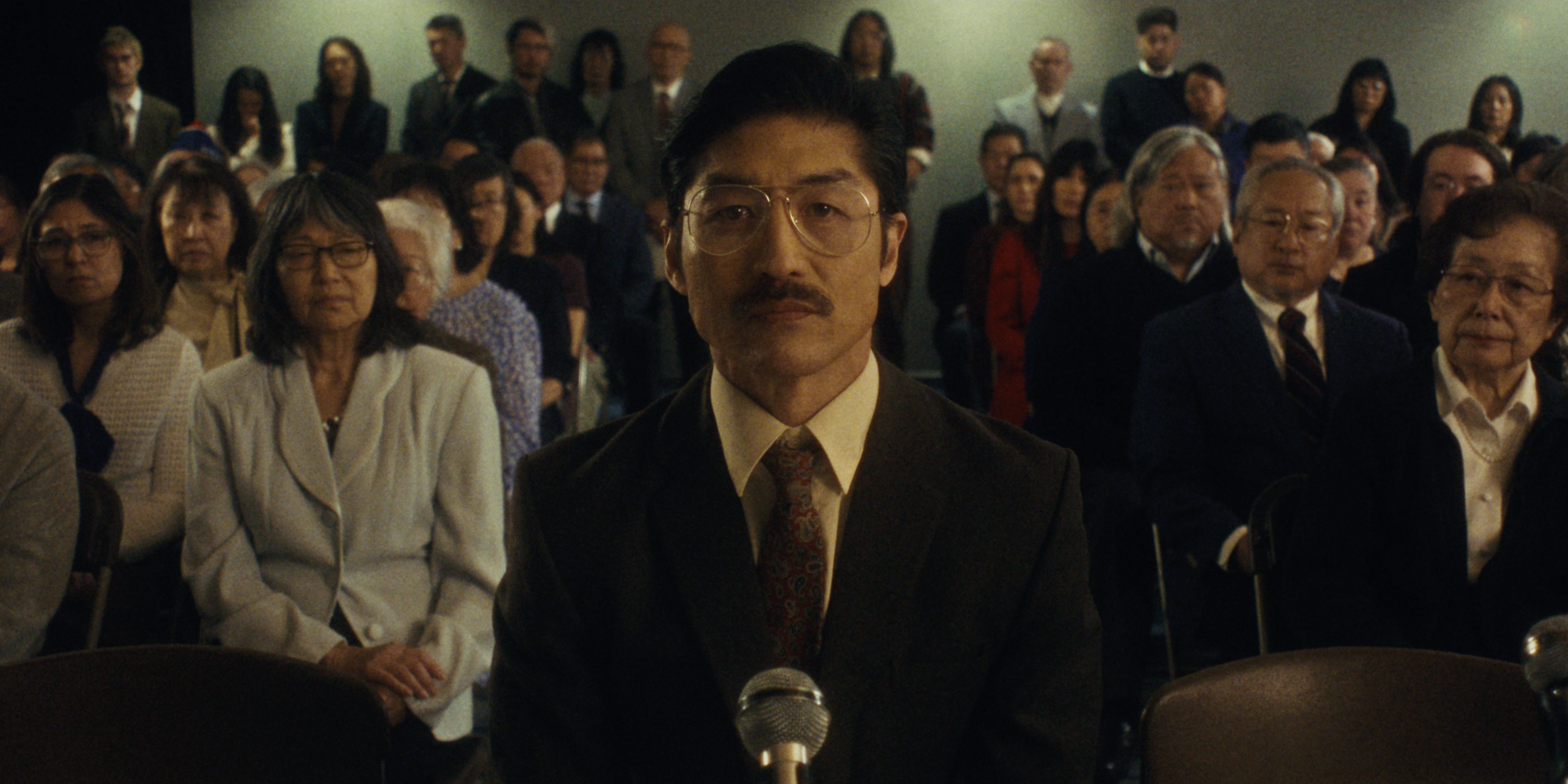 A man with glasses and a mustache in a suit sits in front of a microphone, surrounded by an audience in a conference room.