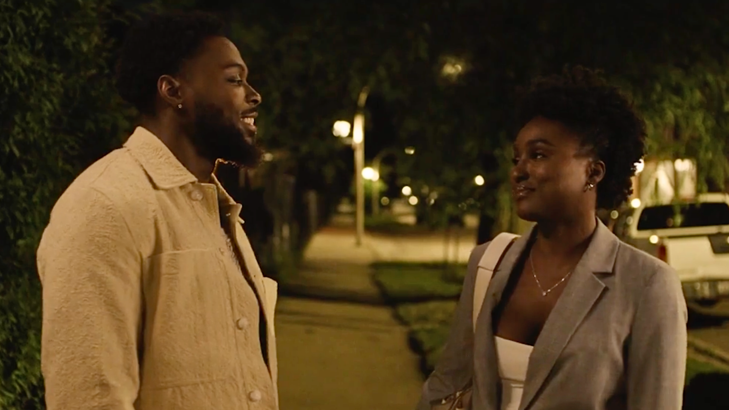 A man and woman are standing on a sidewalk at night, having a conversation. The man is wearing a beige jacket and the woman is in a light gray blazer with a white top. They are outdoors with trees and parked cars in the background, illuminated by streetlights.