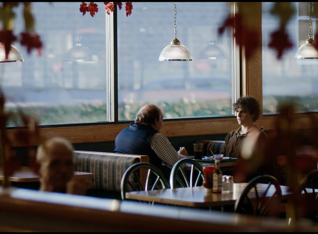 Two people sitting in a cozy restaurant near a large window, engaging in a conversation. The background shows a blurred cityscape outside. The restaurant has warm lighting and wooden accents.