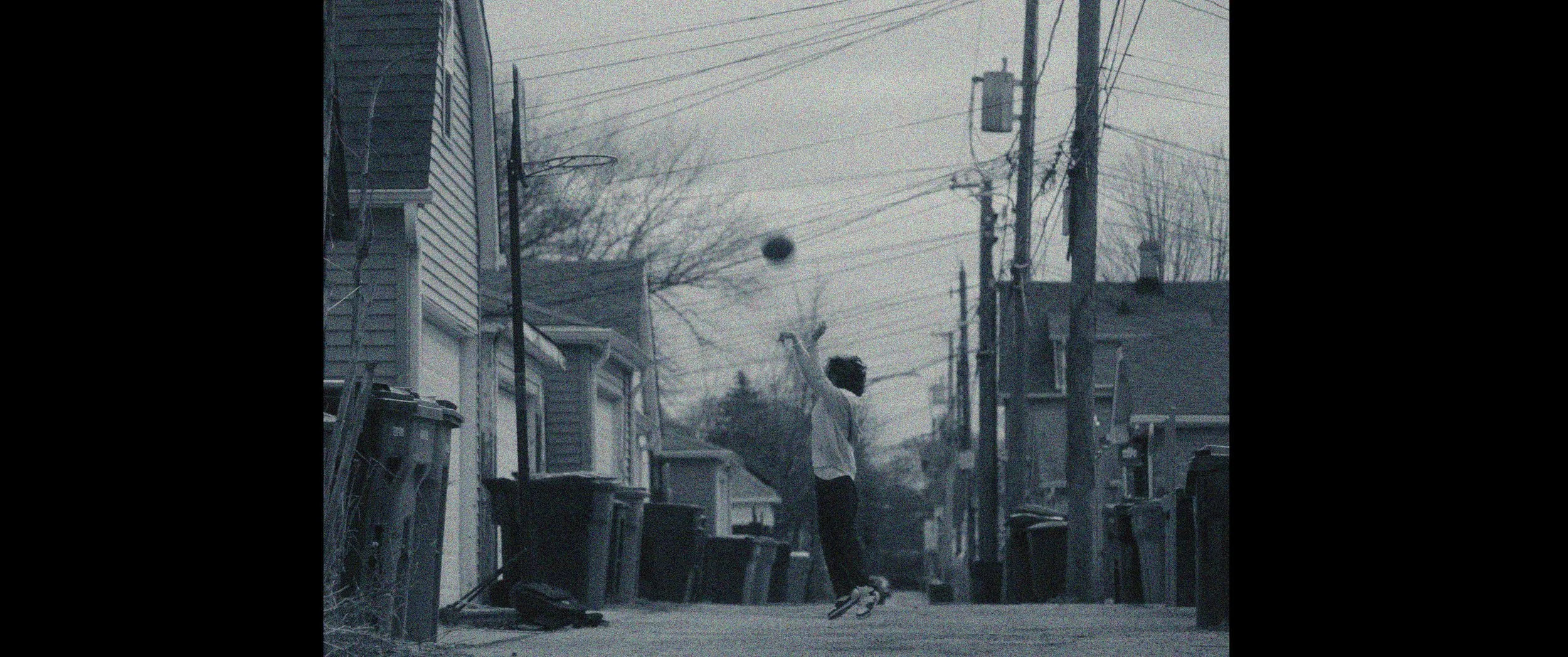 A child in a hoodie and sneakers standing on a deserted alleyway surrounded by houses with trash bins along the sidewalk, with power lines overhead on a foggy day.