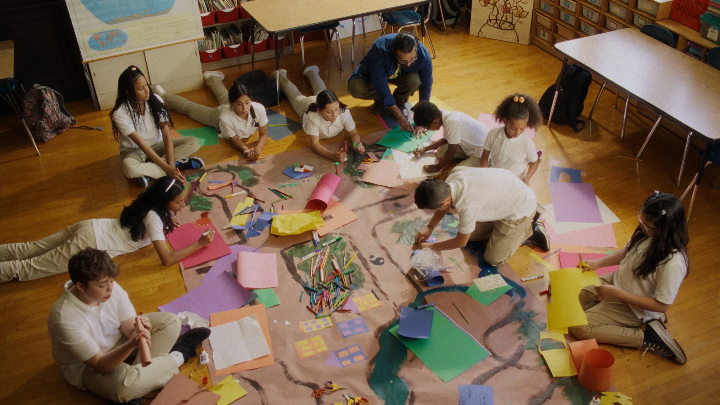 Students and teachers working together on an art project on a large floor mural, surrounded by colored paper, markers, and craft supplies.