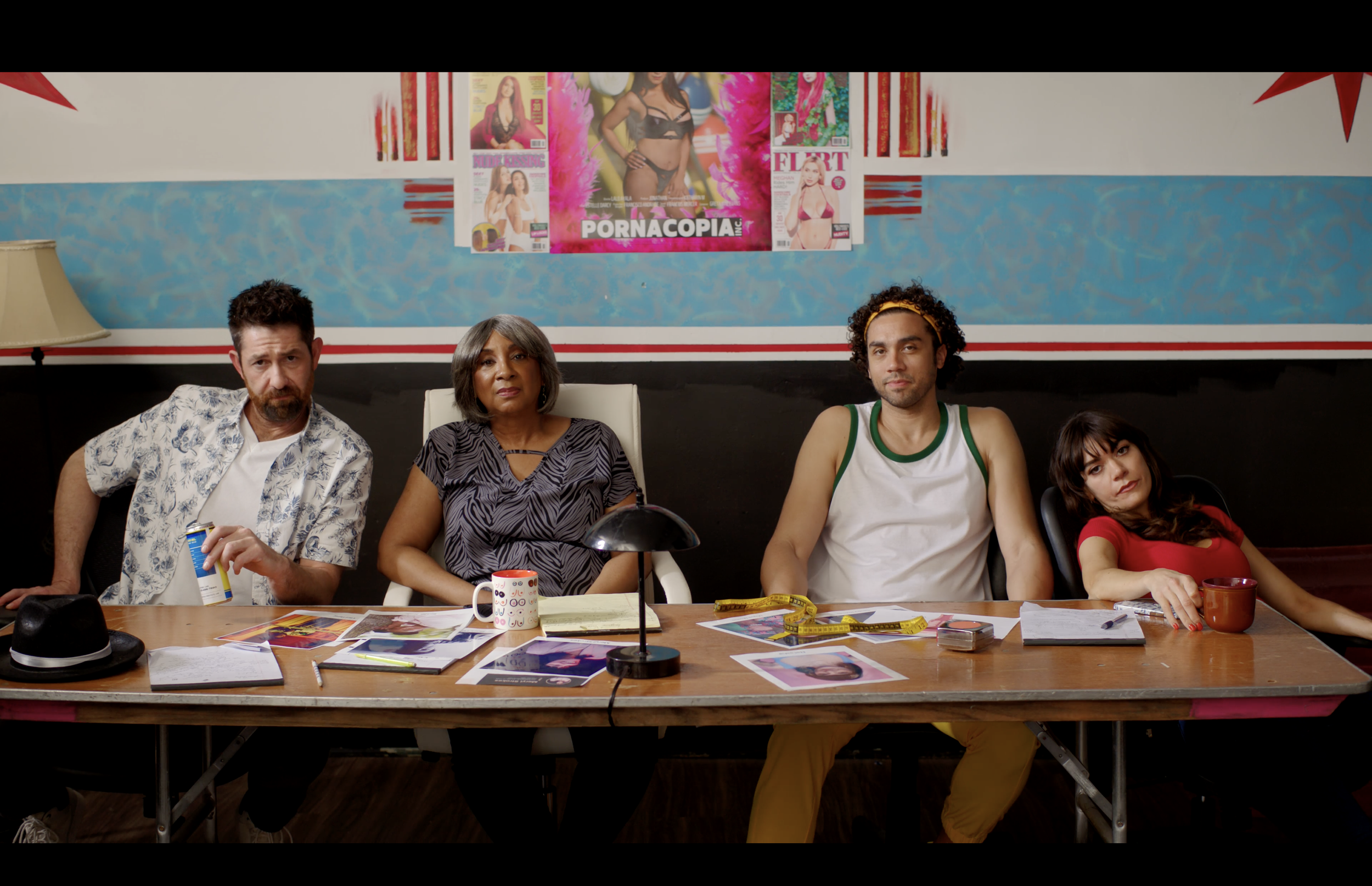 Four people sitting at a cluttered table in a room with colorful posters on the wall, including adult magazine covers. The group appears disinterested or bored.