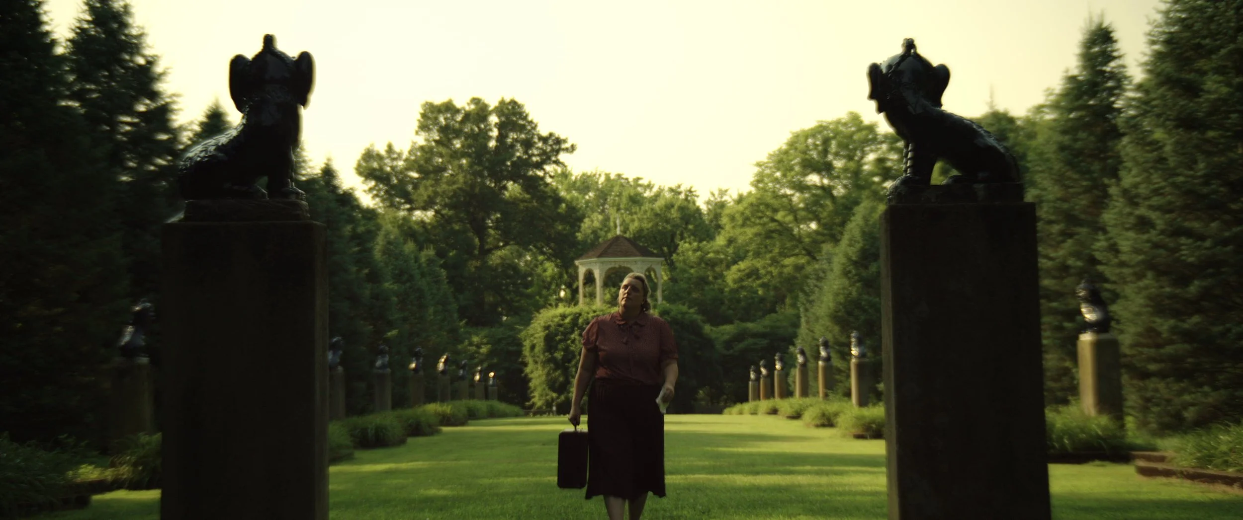 A woman walking in a park-like setting with green grass, trees, and a gazebo in the background, carrying a suitcase.