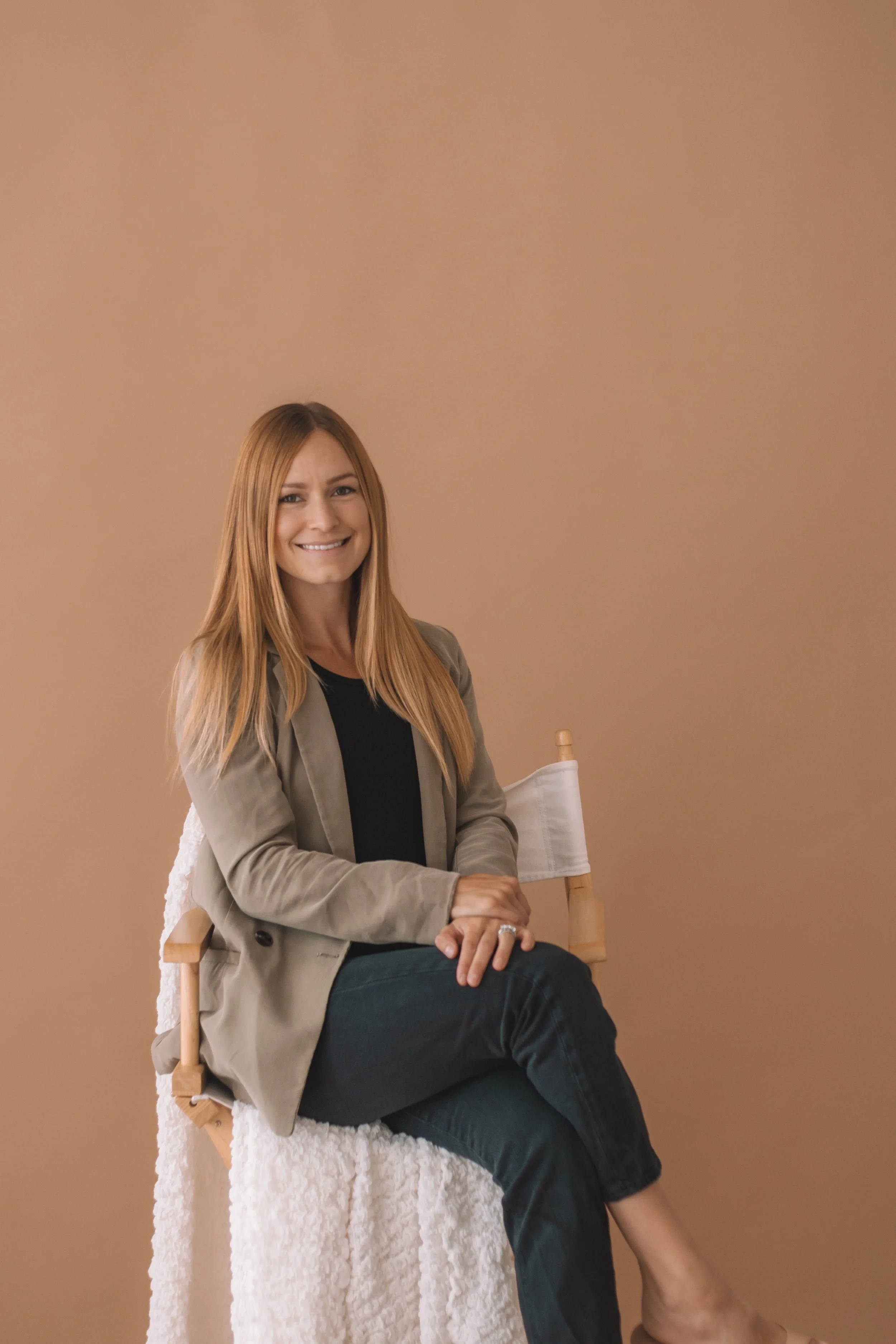 A woman with long blonde hair sitting on a wooden chair with a white cloth draped over the back, smiling at the camera, against a plain beige wall.