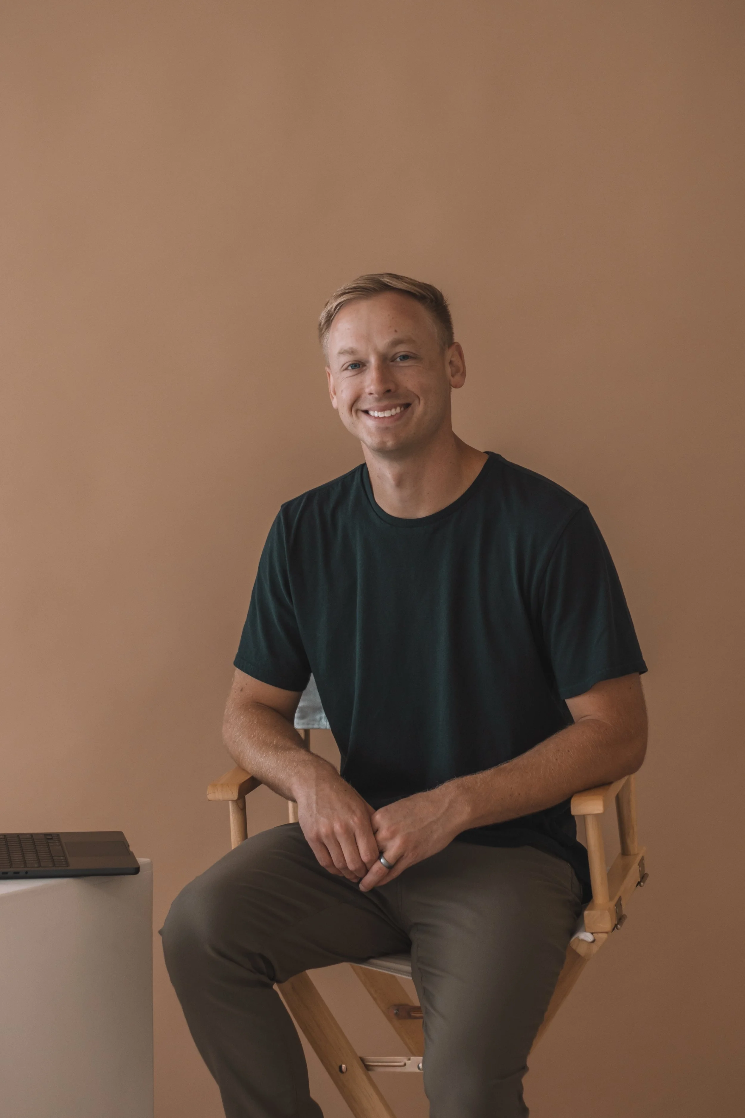 A young man with short blonde hair and a smile, wearing a black t-shirt and gray pants, sitting on a wooden director's chair against a plain brown wall, with a laptop on a white side table nearby.