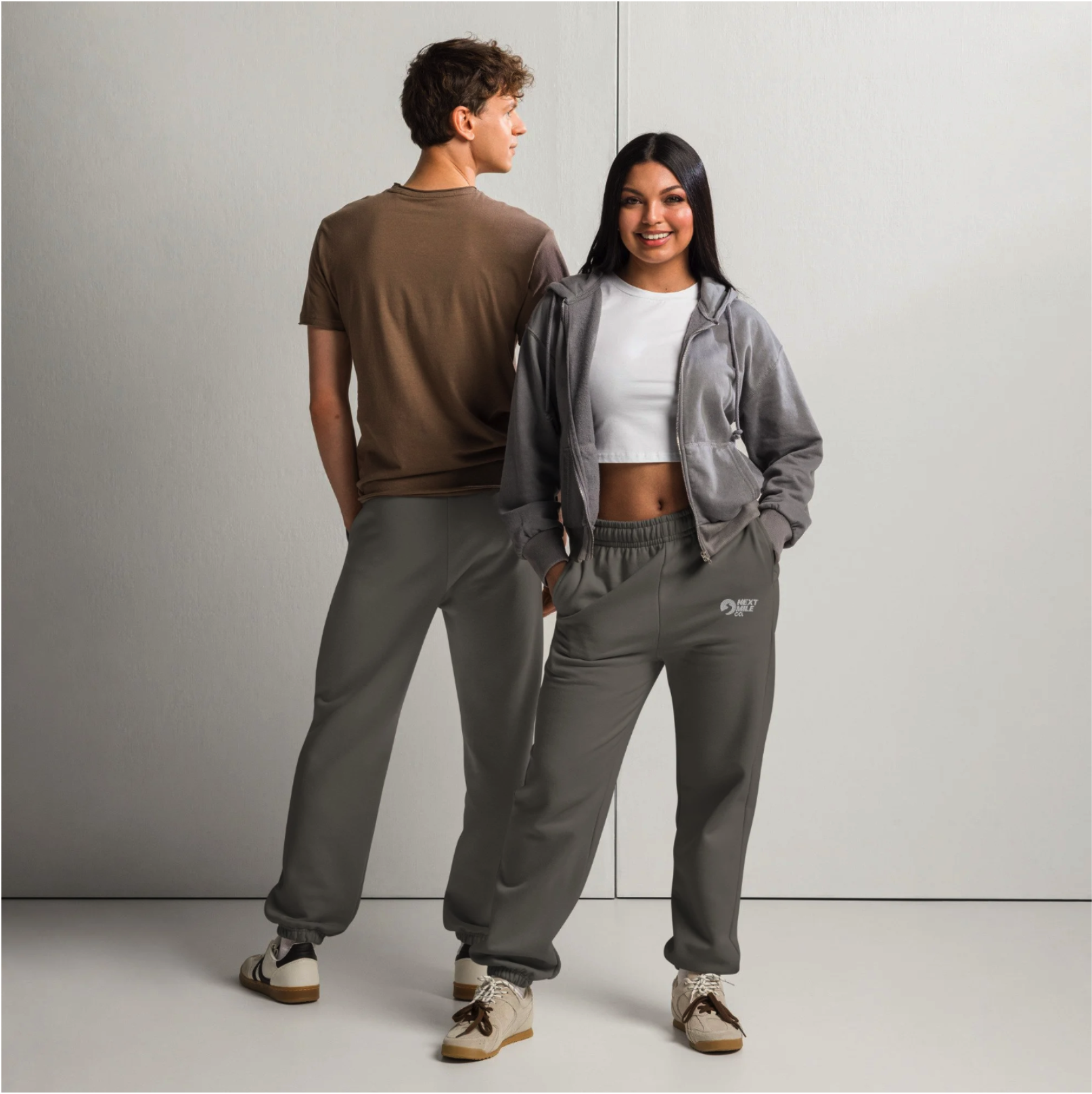 A young man and woman standing against a plain wall, both wearing casual streetwear. The man has curly brown hair, a brown T-shirt, and gray sweatpants, while the woman has long dark hair, a white crop top, a gray zip-up hoodie, and matching sweatpants with sneakers.