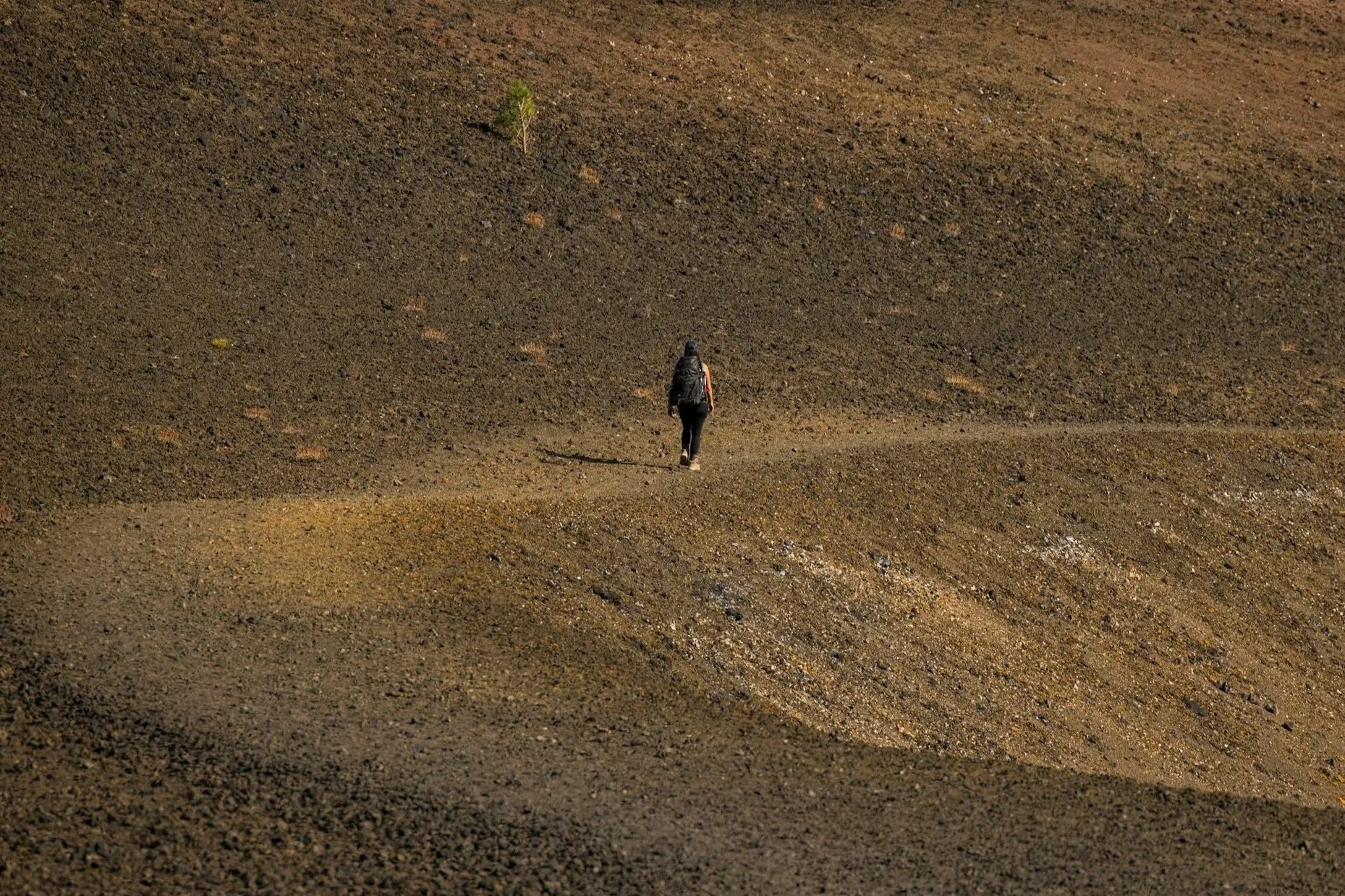 A solitary person with a backpack walking on a wide, barren, uneven dirt landscape with small patches of greenery.