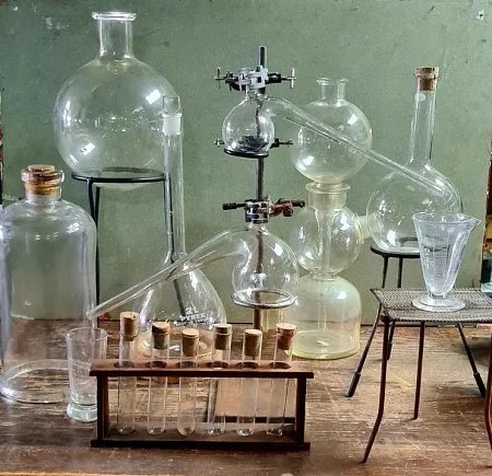 Multiple glass laboratory flasks and test tubes arranged on a wooden table and stands, with some connected by glass tubing, set against a green wall in a chemistry lab.