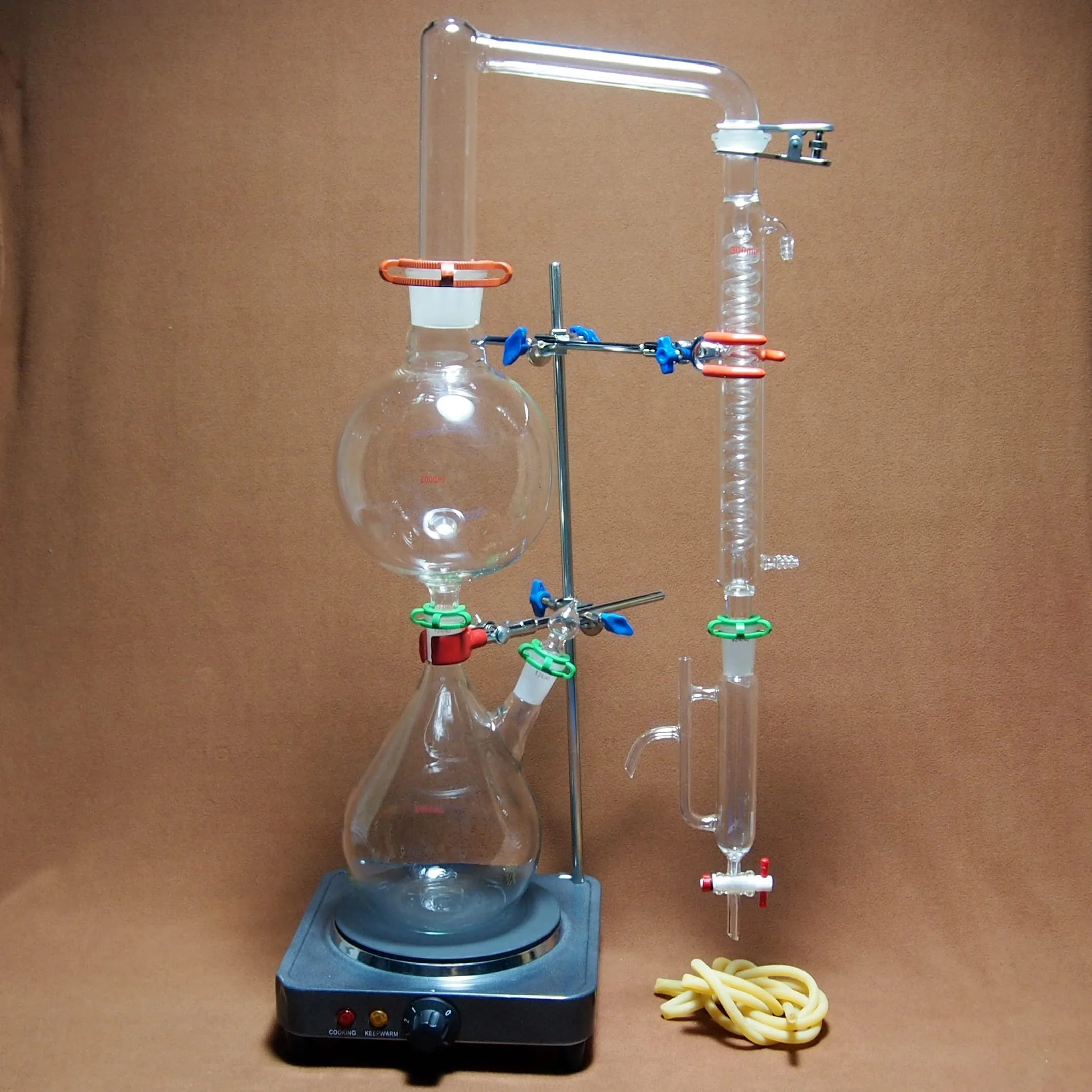 Laboratory glassware setup including a round-bottom flask, a condenser, and connecting glass tubes secured with colorful clamps, placed on a heating plate with a power switch and control knob. A yellow rubber tubing is coiled next to the apparatus.