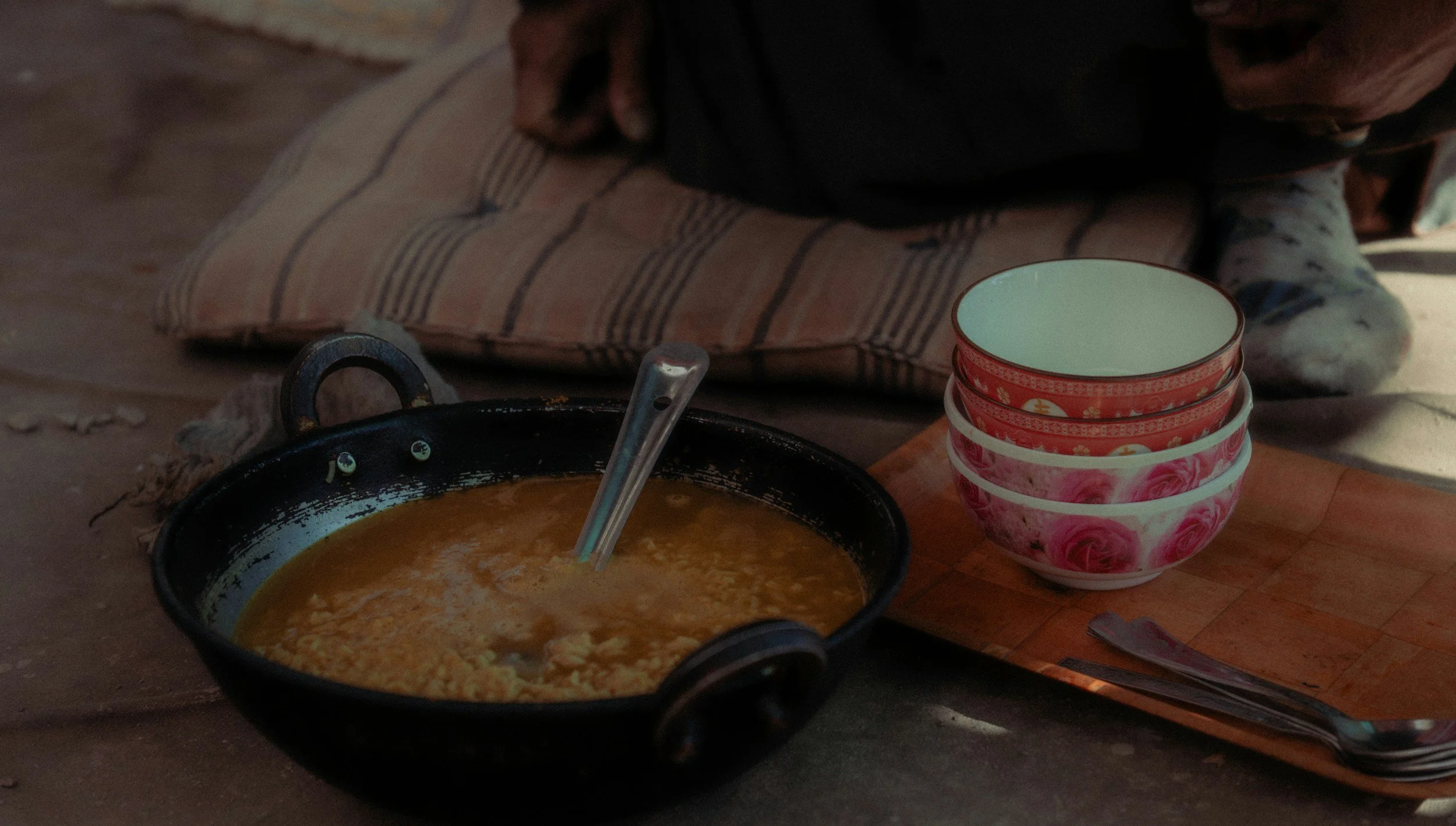 A bowl of soup with a spoon on a tray, and four stacked bowls with pink floral designs next to it, set on a wooden surface with a person sitting in the background.