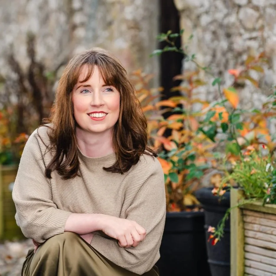 A woman with shoulder-length brown hair and blue eyes smiling outdoors in front of autumn leaves and potted plants.
