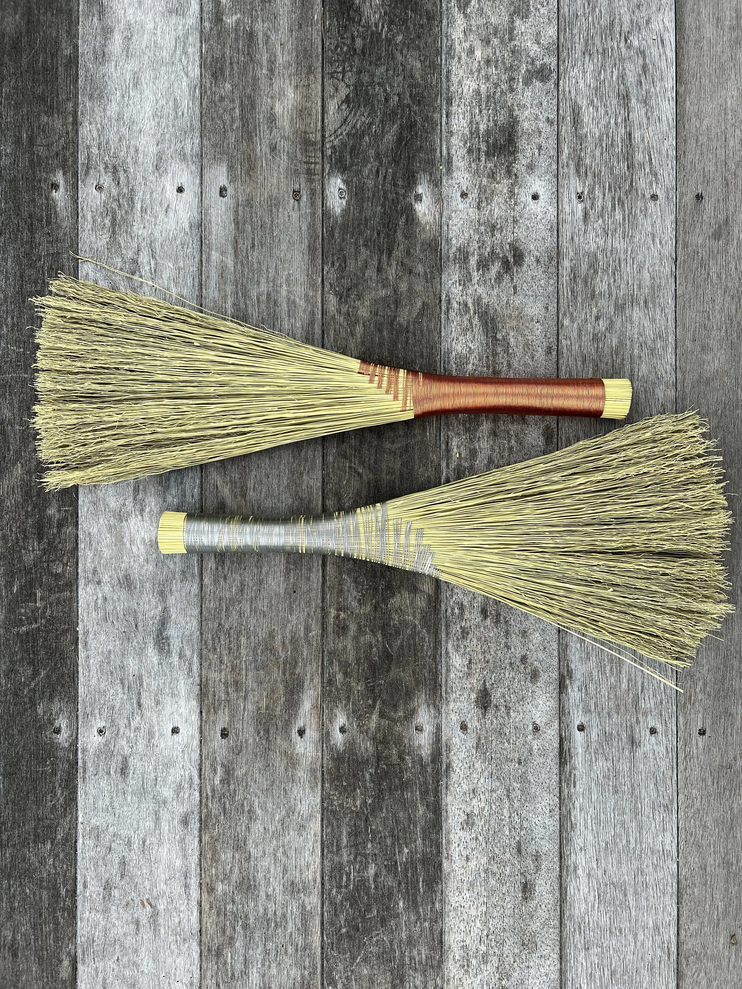 Two traditional straw brooms with woven handles, placed on a weathered wooden surface.