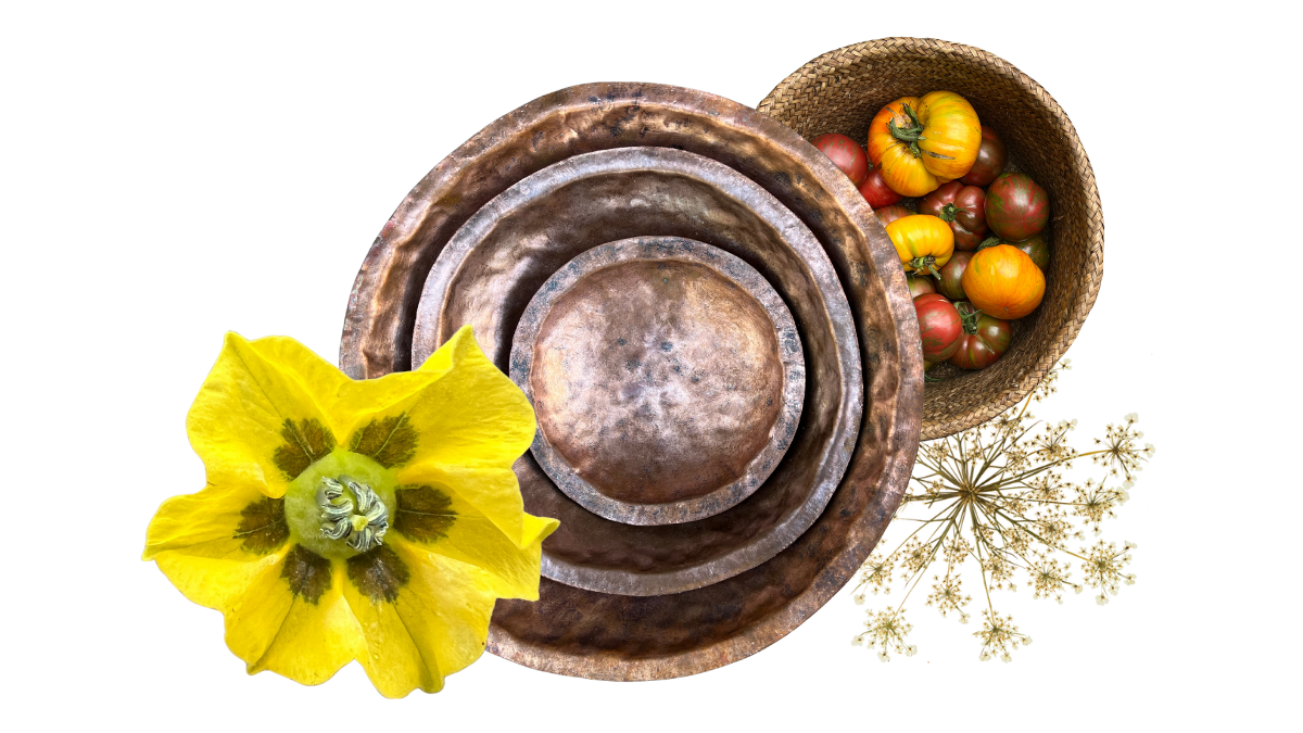 Three concentric metal bowls, a yellow flower, a basket of heirloom tomatoes, and a sprig of white flowers arranged on a surface.