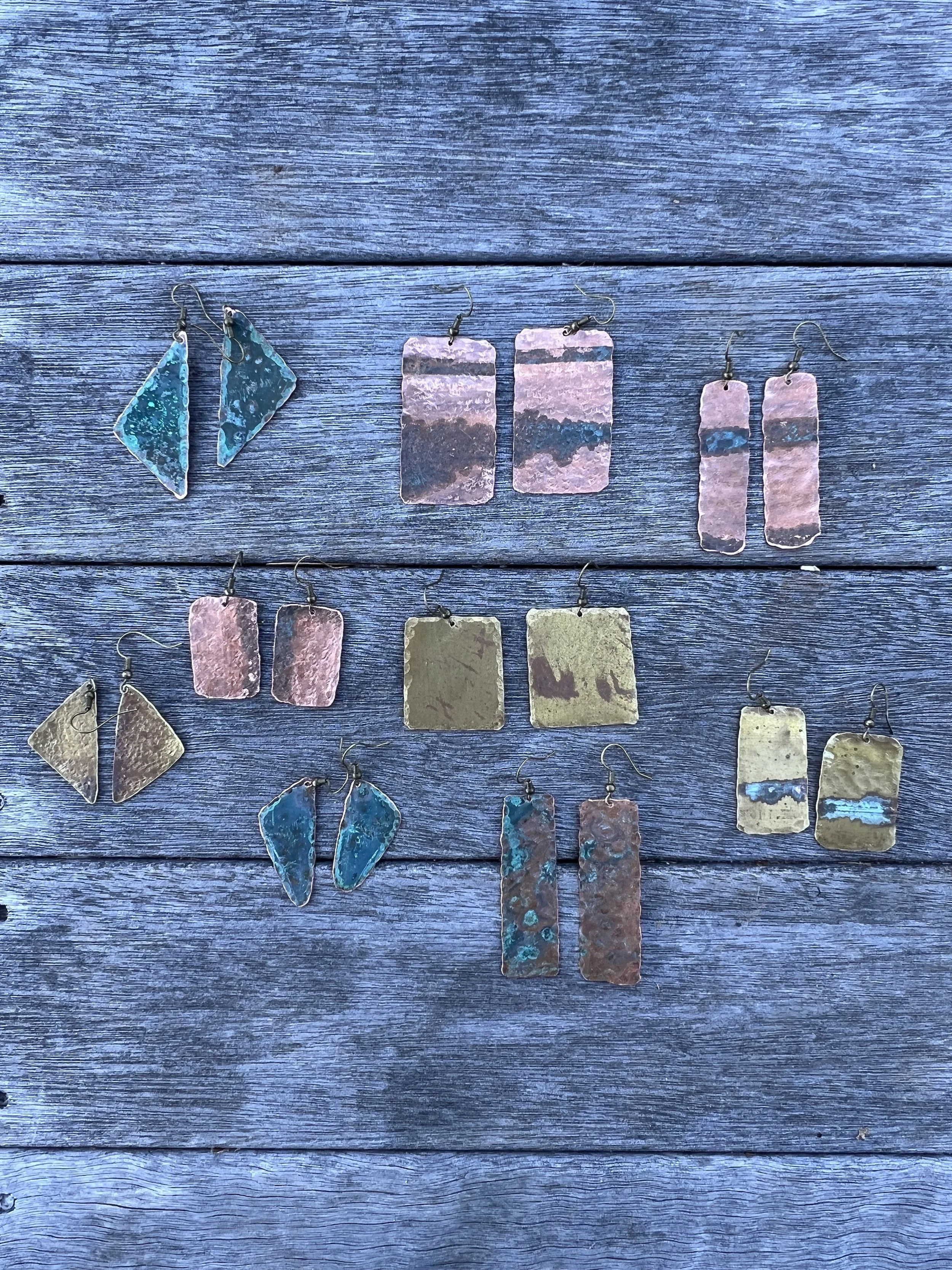 Collection of handmade metal earrings with various rectangular and triangular shapes, displaying a range of oxidized copper and brass finishes, arranged on a weathered wooden surface.
