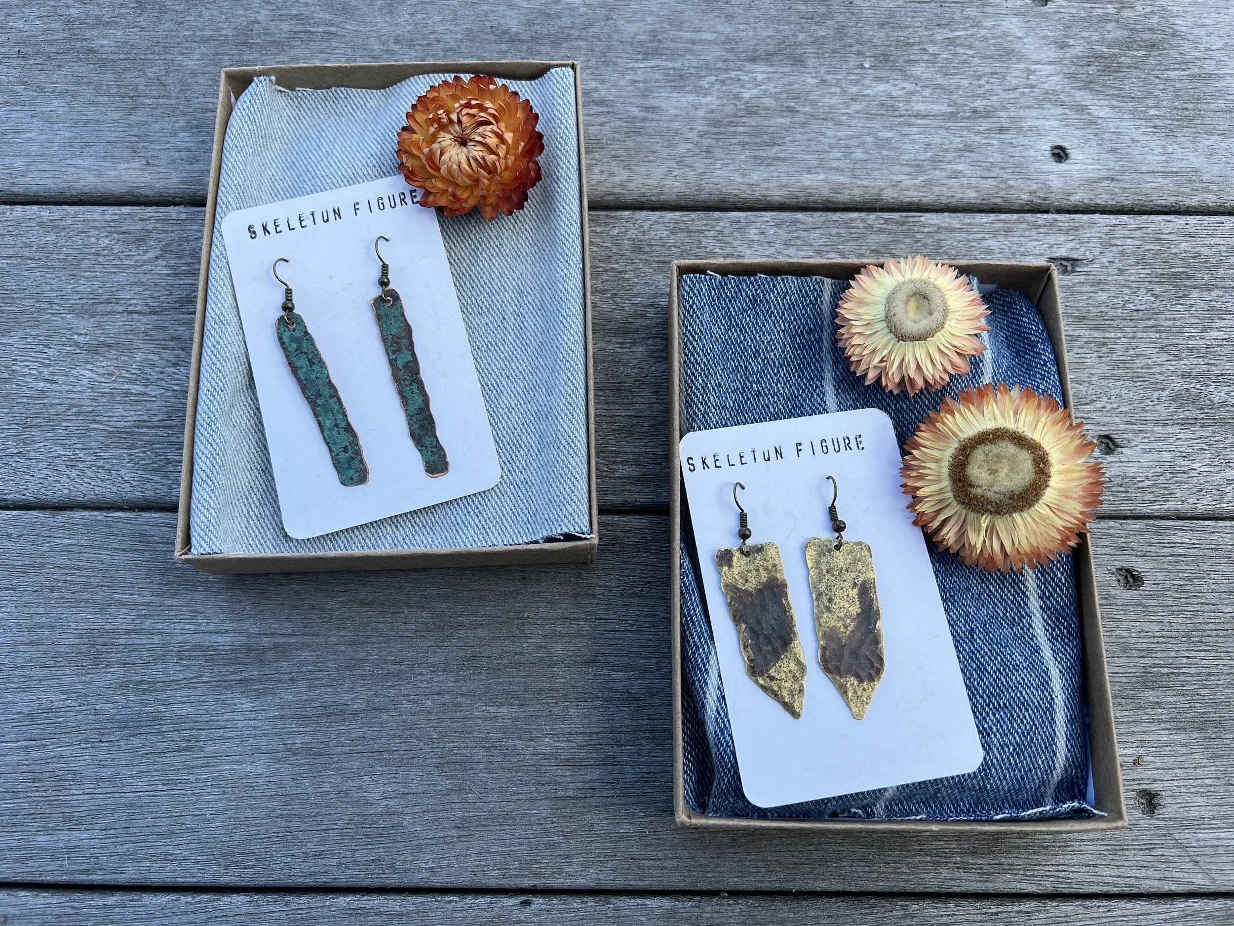Two boxes of handmade earrings, one with turquoise-colored elongated rectangular earrings and the other with gold-colored irregular shaped earrings, each box decorated with dried flowers on a wooden surface.