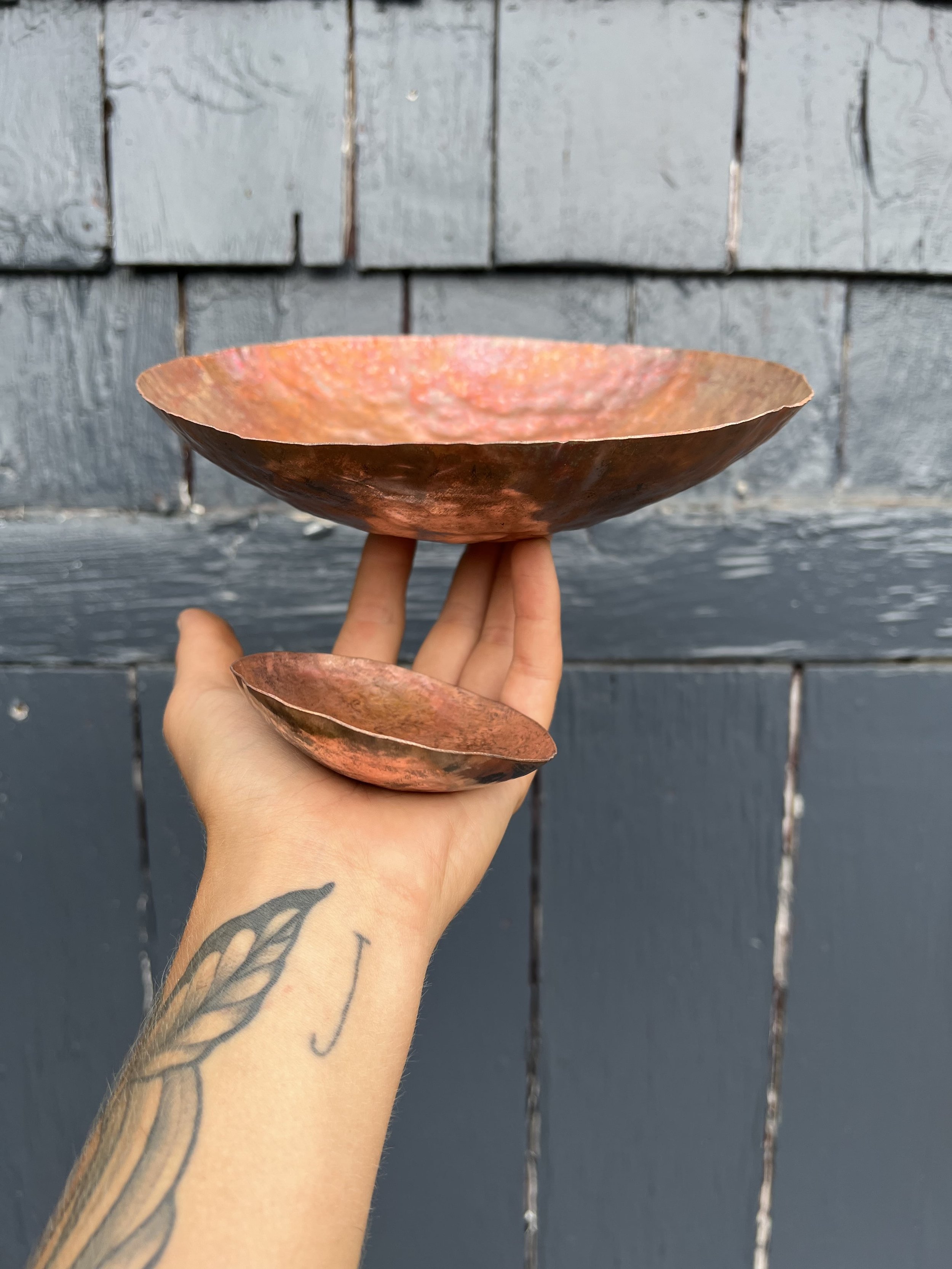 A person with a tattoo on their forearm holding two rust-colored metal bowls, one larger and one smaller, in front of a weathered gray wooden wall.