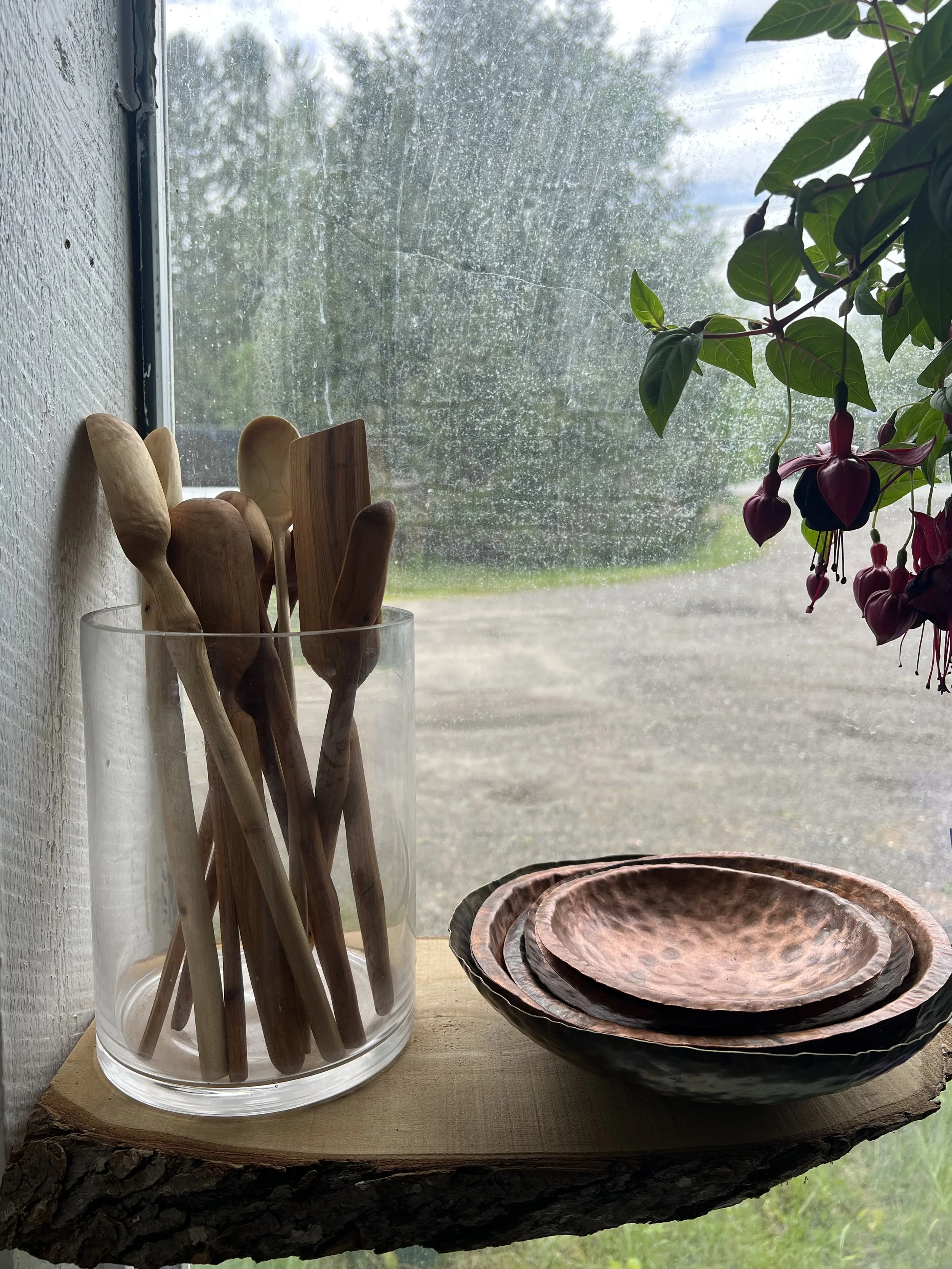 Wooden kitchen utensils in a clear glass container, stacked ceramic bowls with a textured finish, on a rustic wood shelf by a window with a rain-streaked glass, next to green leaves and hanging fuchsia flowers outside.