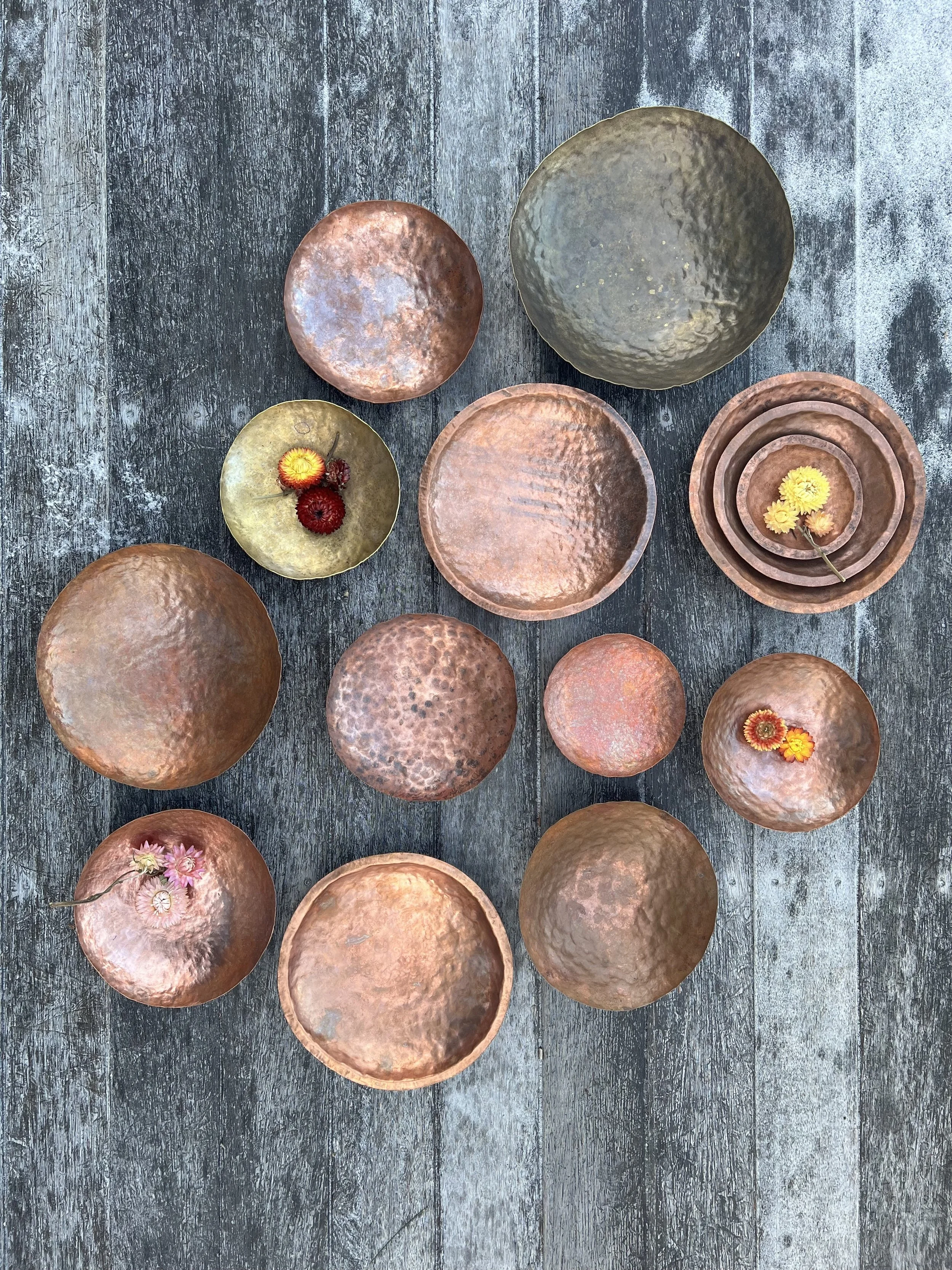 Set of 12 textured copper and metal bowls with flowers on rustic wooden surface.