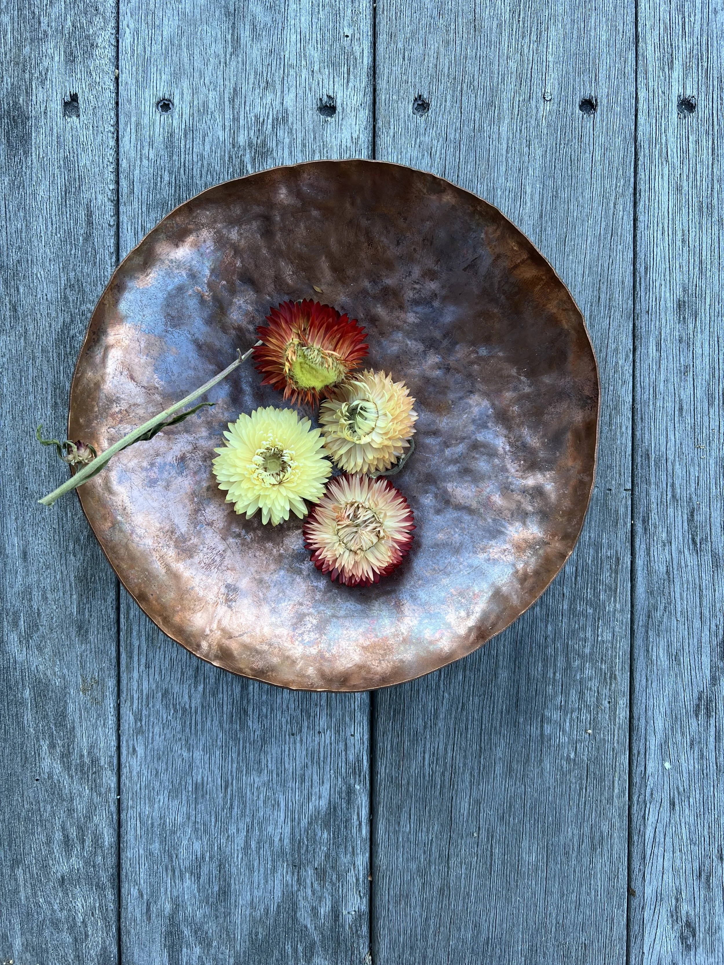 A round, textured copper plate holding five colorful flowers, placed on a weathered blue wooden surface.