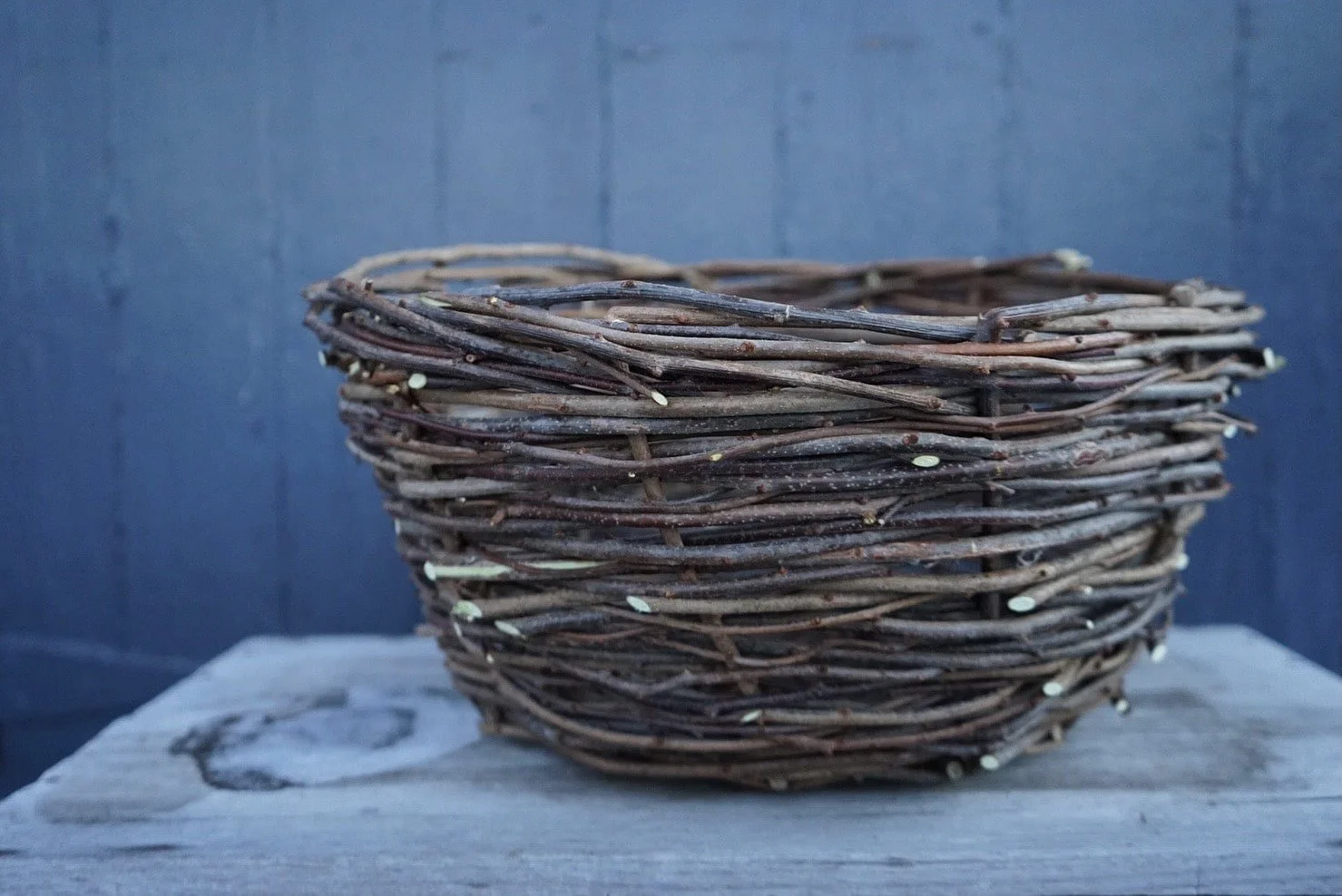 A woven bird's nest made of thin, dark brown twigs placed on a weathered wooden surface with gray background.