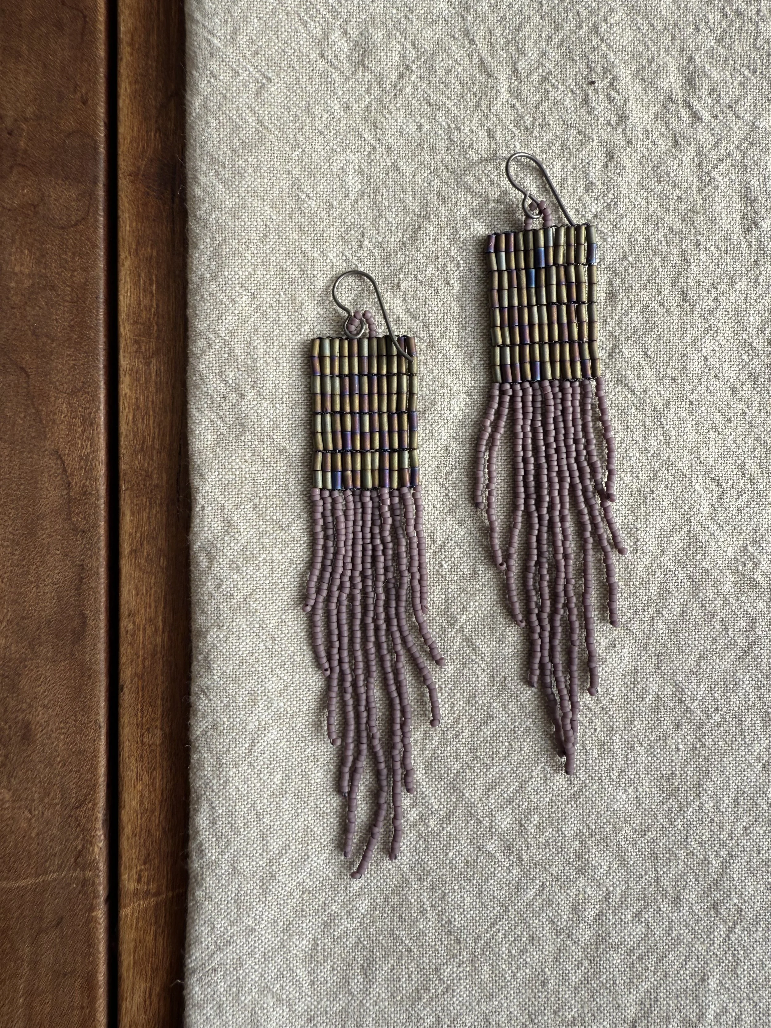 Pair of beaded earrings with square top sections and long hanging fringe made of small pink and metallic beads, placed on a beige textured fabric surface.