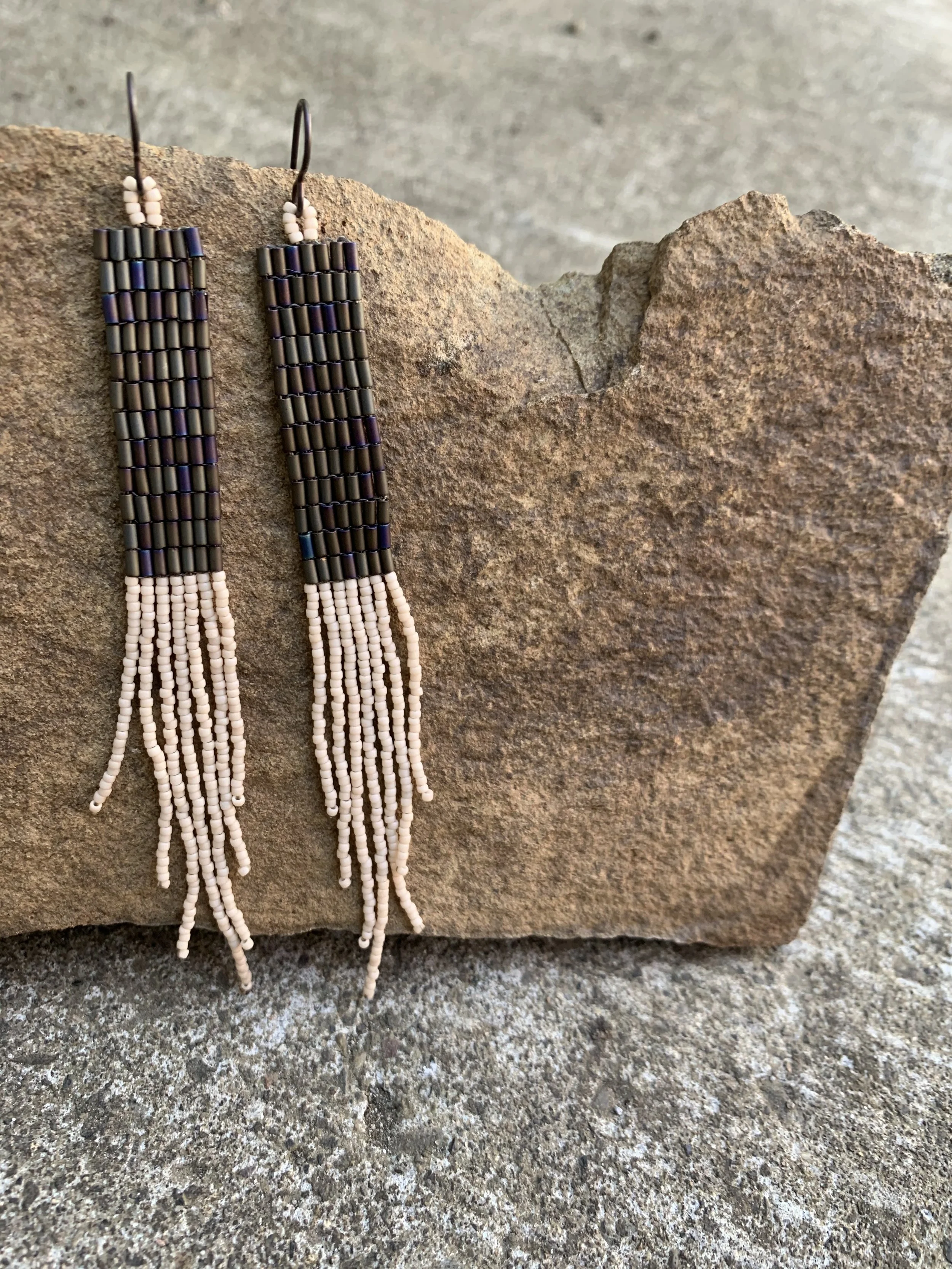 Pair of beaded earrings with black, purple, and white beads, hanging from a piece of rock.