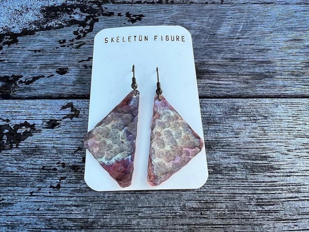 The image shows a pair of earrings with large, irregularly shaped, metallic-colored pieces, hanging on a white card labeled 'Skeleton Figure.' The background surface is weathered, grayish wood.