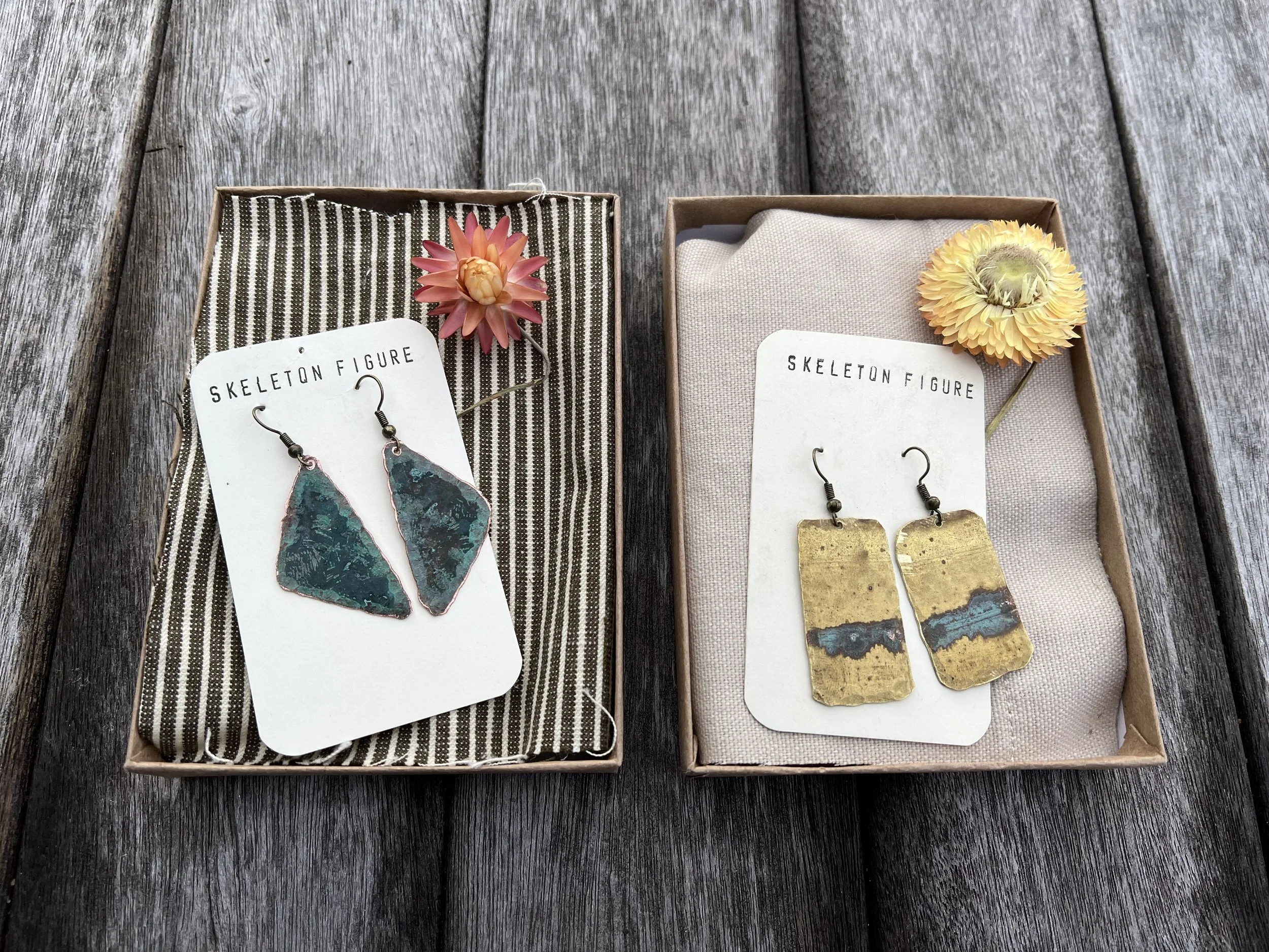 Two boxes of earrings with flowers on a wooden surface. The left box contains green and black earrings with a pink flower, and the right box contains gold and blue earrings with a yellow flower.