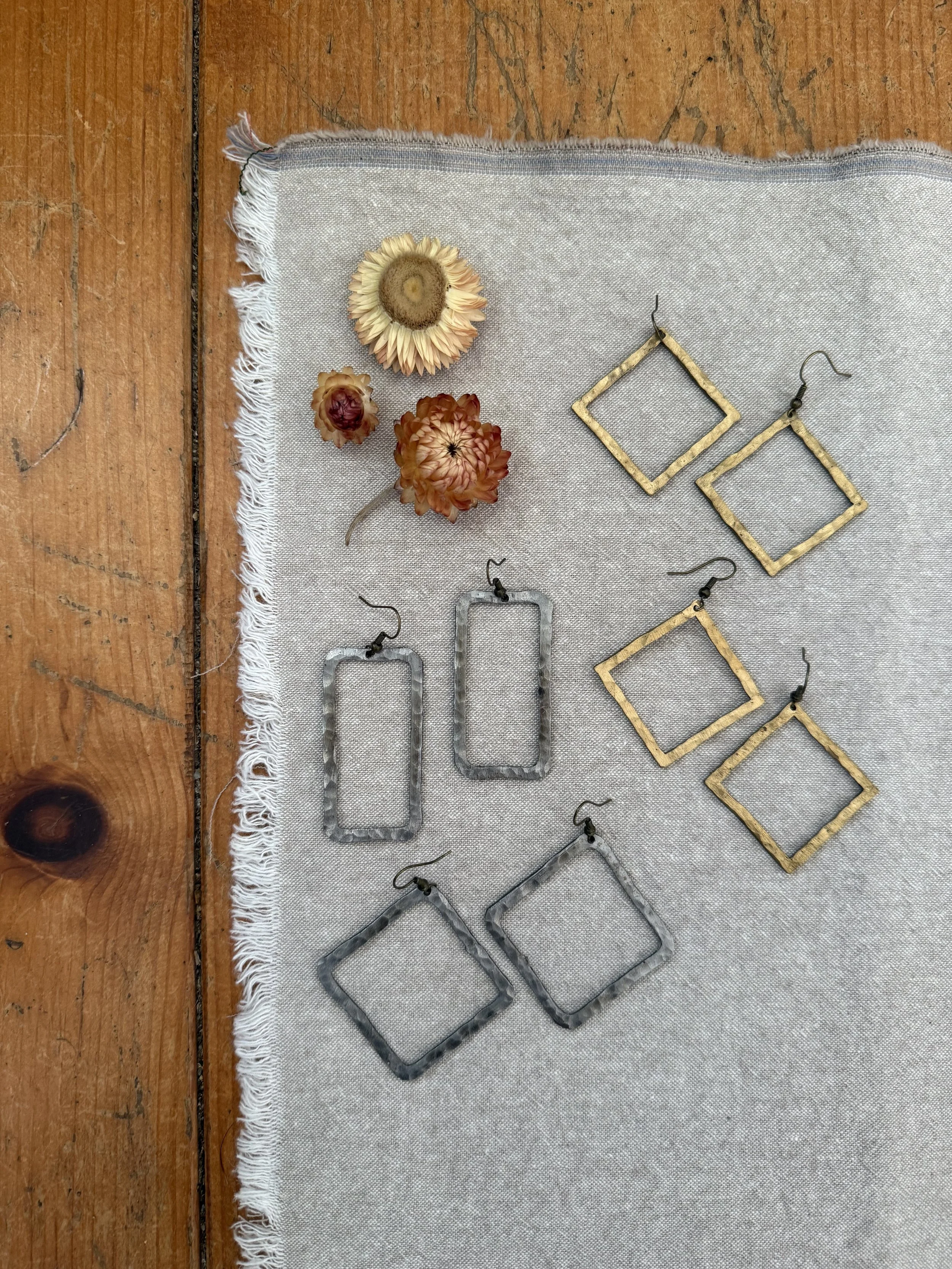 Metal earrings in gold and silver square shapes, placed on a grey textile on a wooden surface, with dried flowers at the top left.