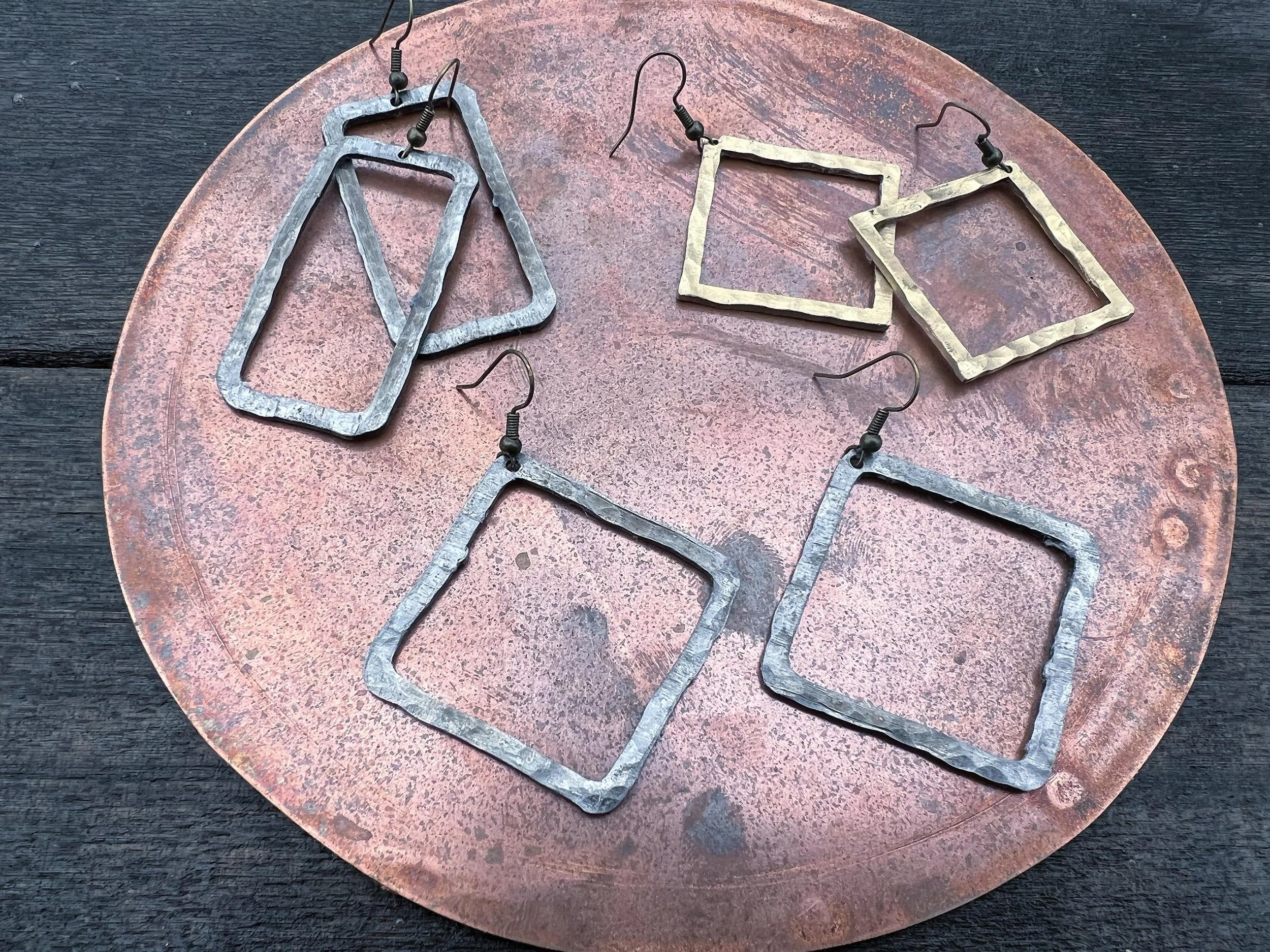 Several pairs of handcrafted metal earrings with rectangular and square shapes are displayed on a round, rusty metal tray.