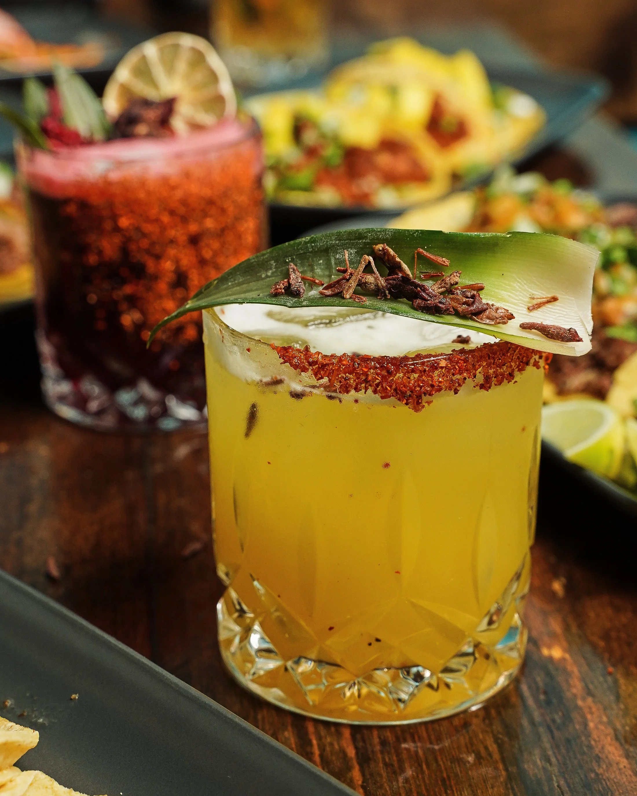 A yellow cocktail garnished with a jalapeño slice, a lime wedge, and a chili powder rim on a textured glass, with a blurred background of colorful dishes and another drink.