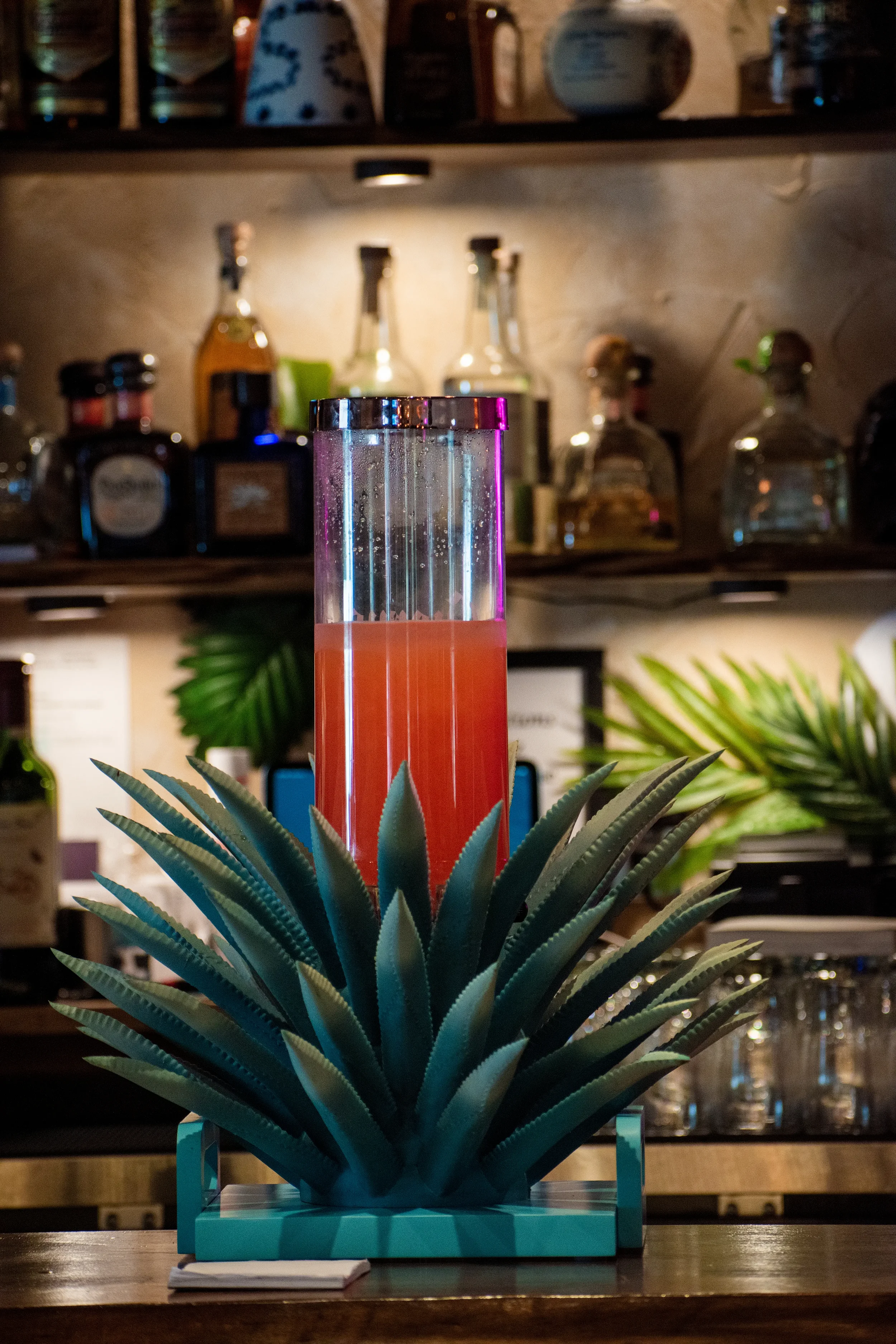 A decorative pineapple-shaped object with green leaves surrounds a tall glass container filled with a pink beverage, set on a bar counter.