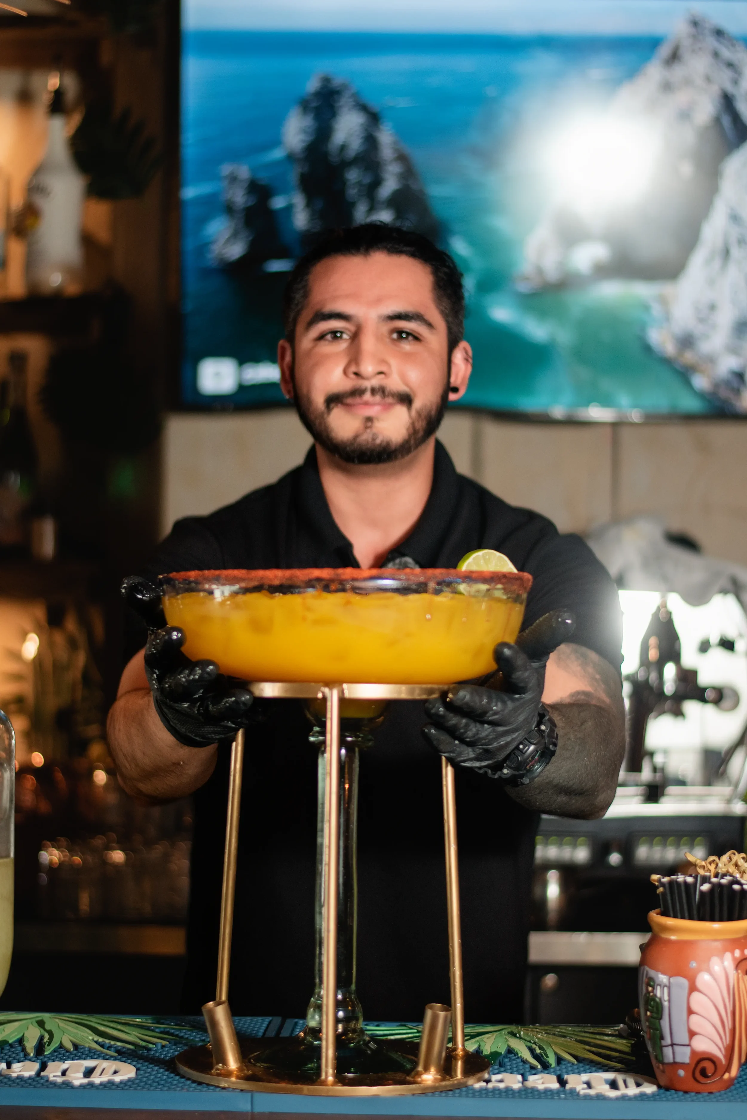 A bartender holding a large bowl-shaped cocktail garnished with a lime wedge, standing behind a bar with a tropical background on a TV screen.