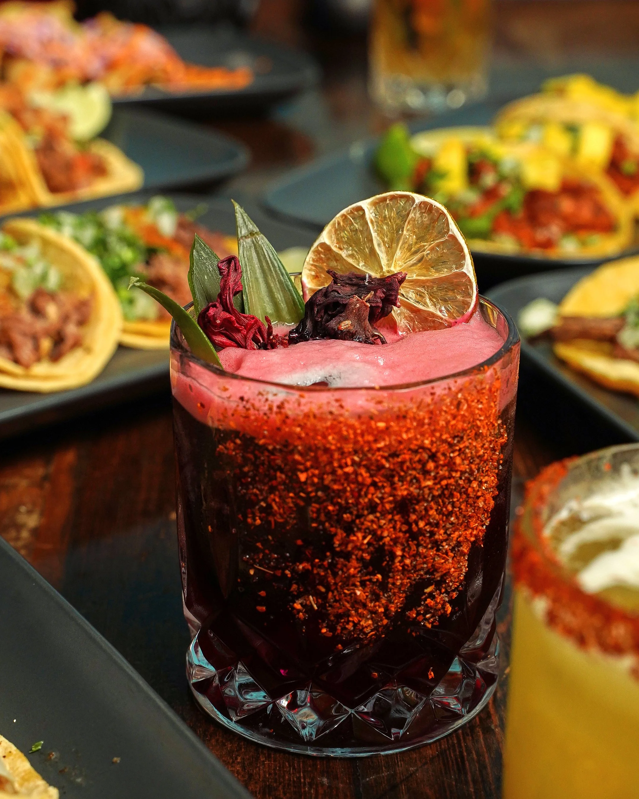 A colorful cocktail garnished with dried lime slice, red hibiscus flower, and a green leaf, with a background of various Mexican dishes including tacos.