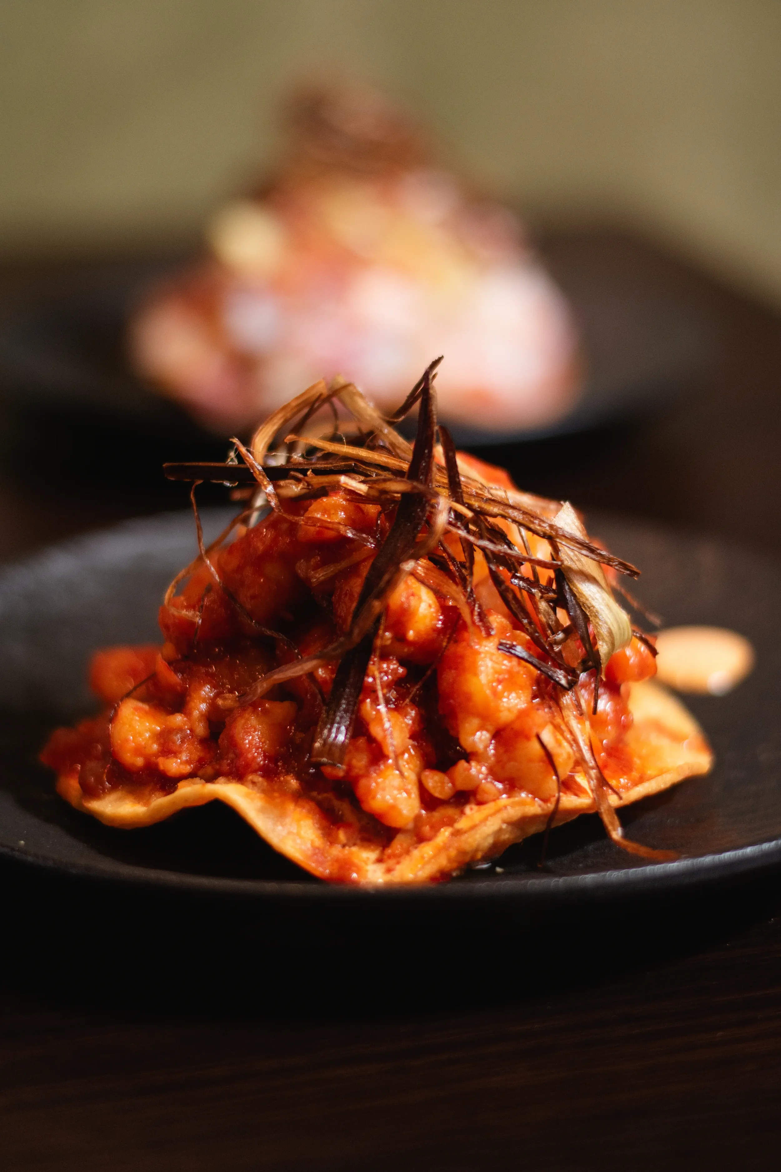 Close-up of a spicy chicken  tostada taco topped with shredded chicken, tomato sauce, and crispy shredded bits on a black plate.