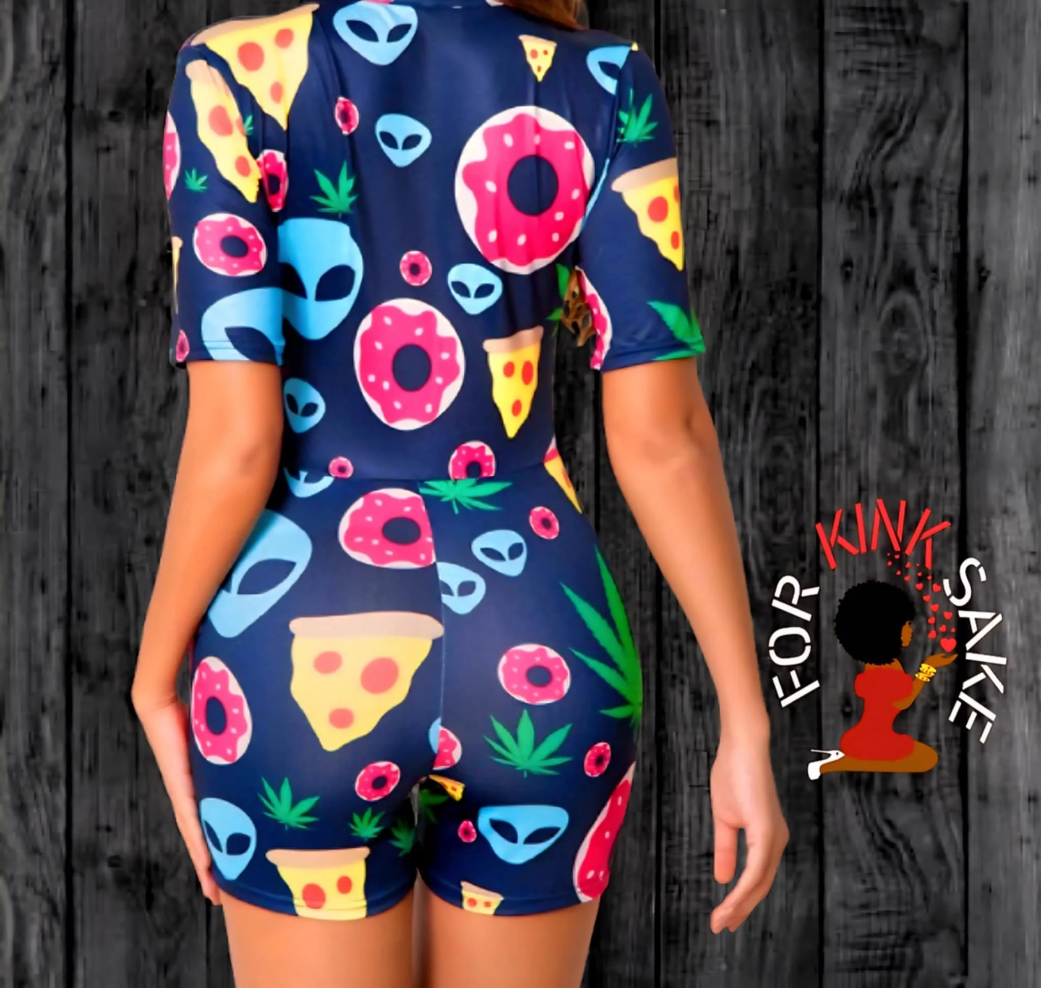 A woman wearing a short-sleeved, form-fitting romper with a dark blue background, decorated with colorful patterns of pizza slices, donuts, alien faces, and green leaves, standing against a black wooden wall.
