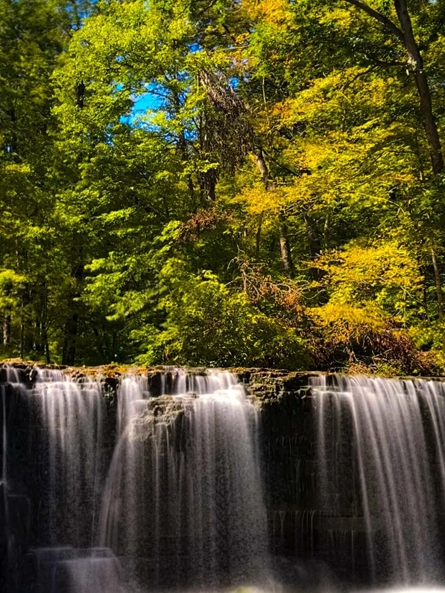 A scenic view of a waterfall flowing over a rocky ledge surrounded by lush green and yellow autumn foliage in a forest.