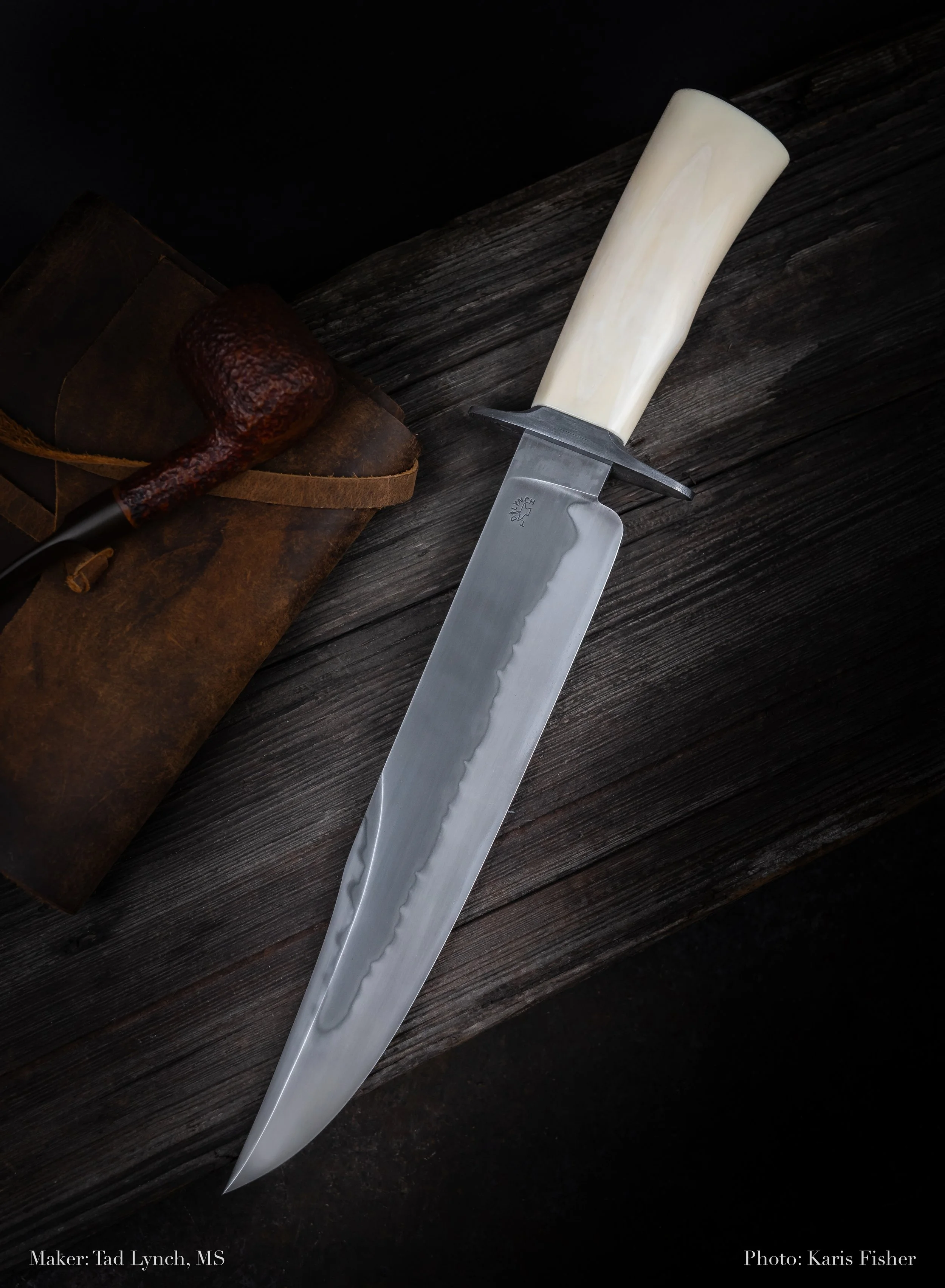 A large fixed-blade knife with a stainless steel blade and a white handle, resting on a dark wooden surface, with a tobacco pipe and a leather pouch nearby.