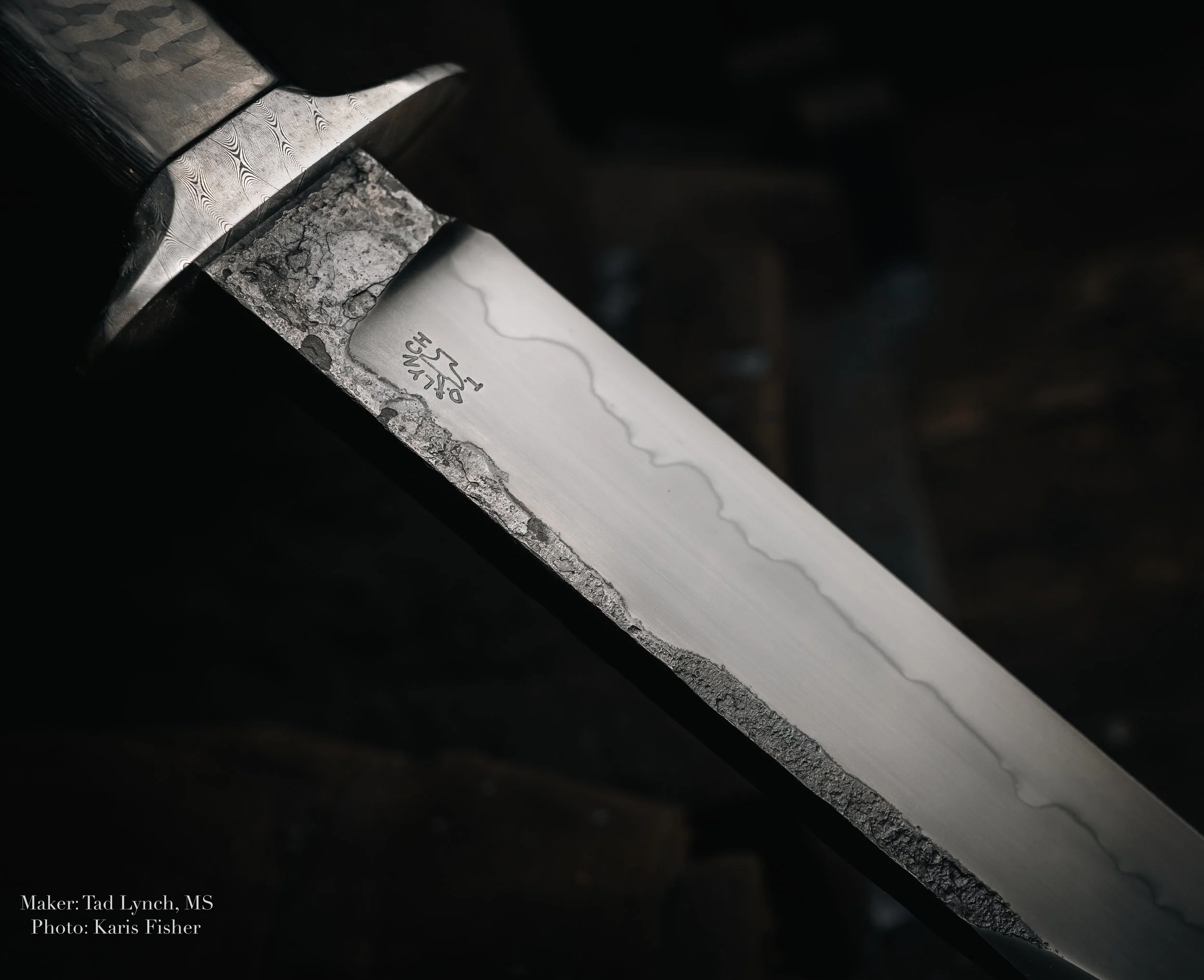 Close-up image of a steel blade with the maker's mark, showing a textured edge and smooth cutting surface, with a dark, blurred background.