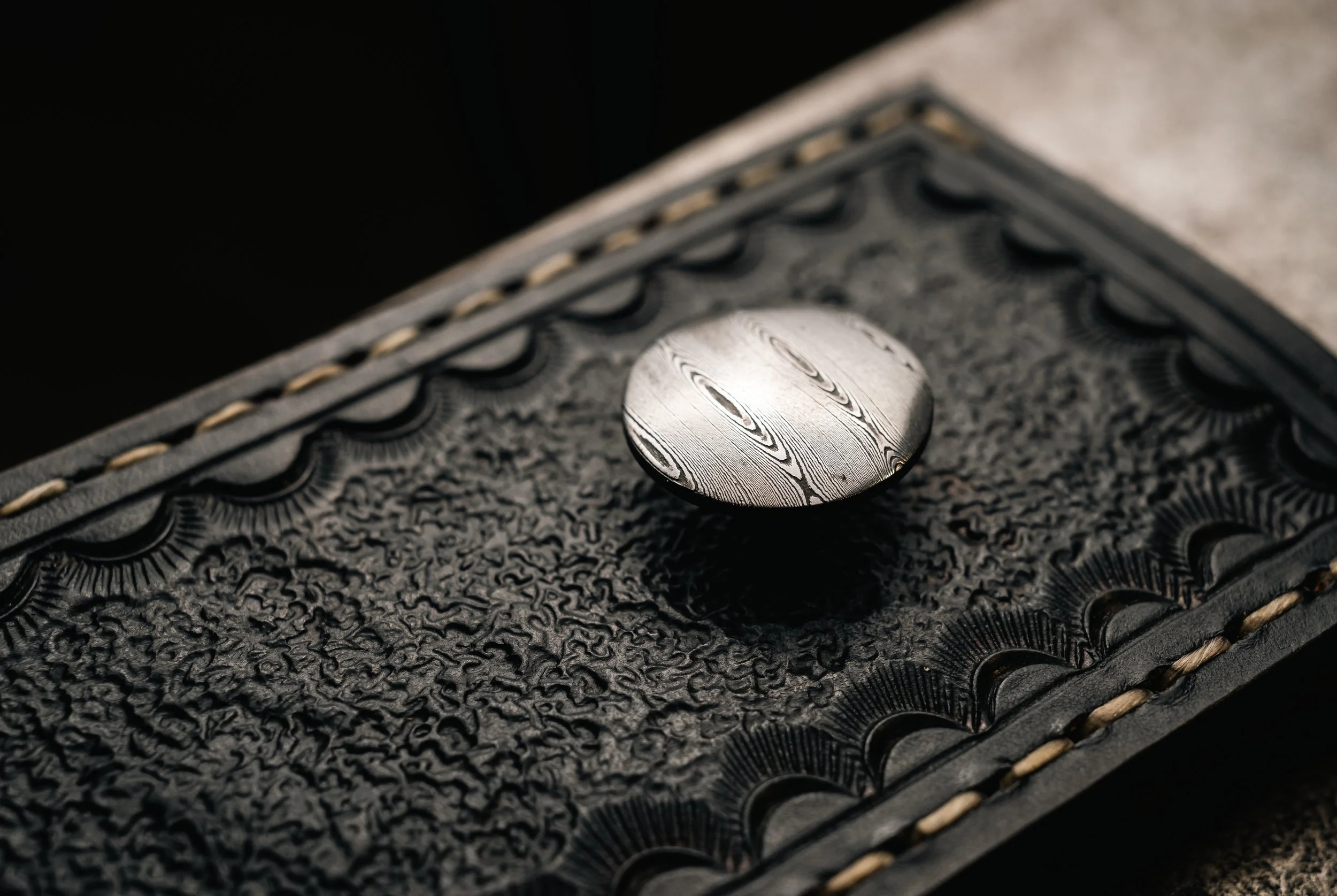 Close-up of a textured leather item with decorative stitching and a metallic button featuring a woodgrain pattern.