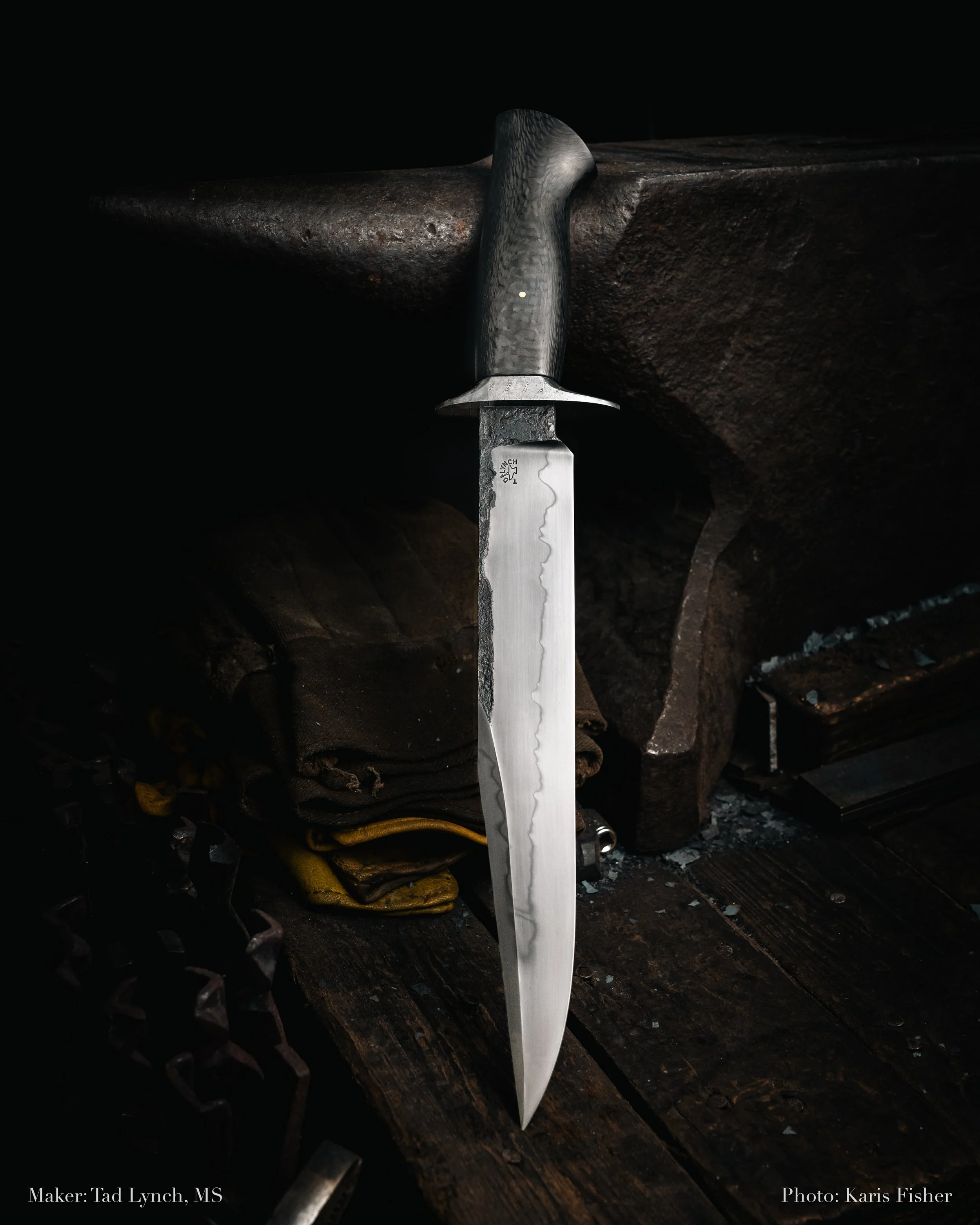 A large handmade knife with a dark textured handle, mounted on an anvil in a blacksmith's forge setting.