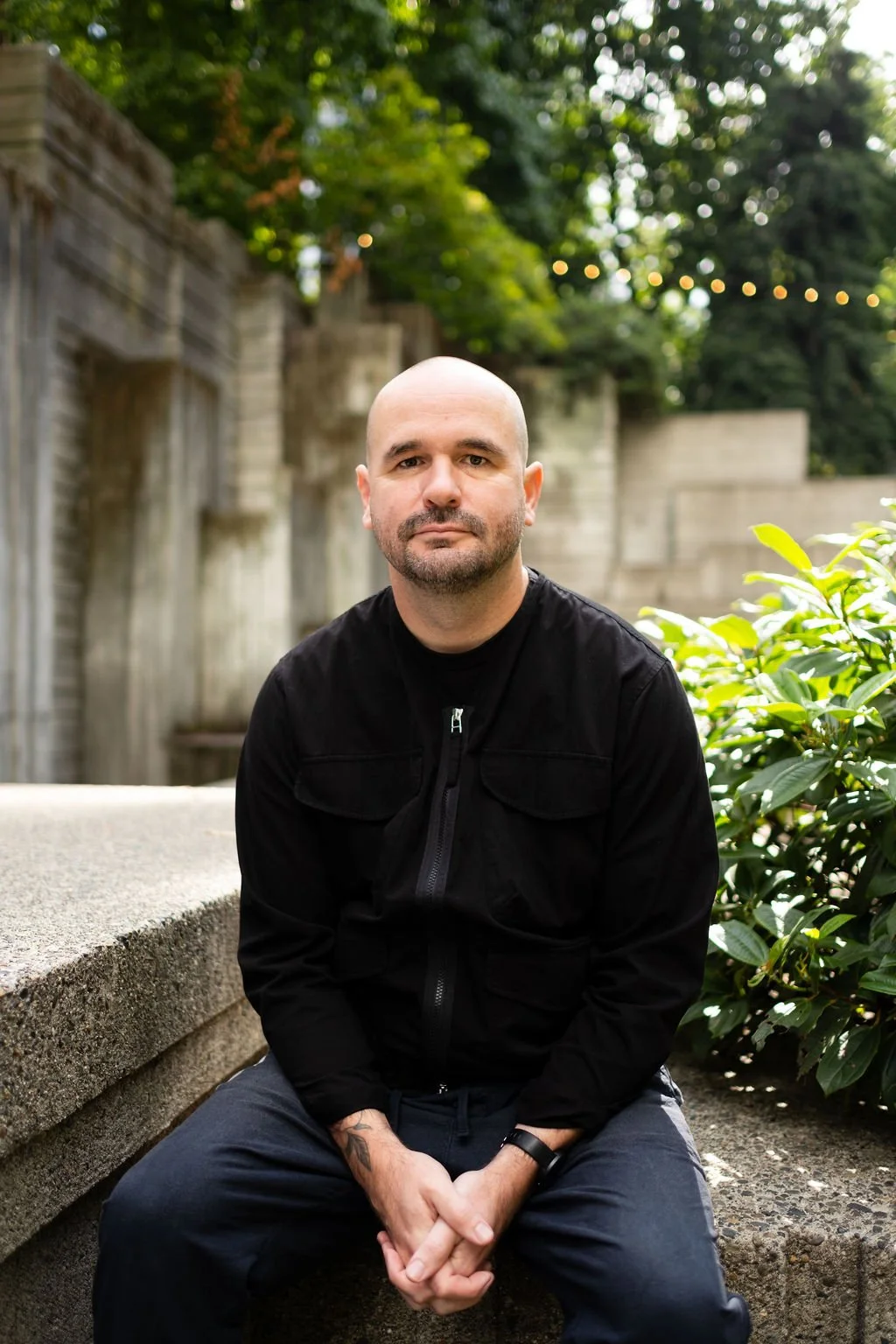 A bald man with a beard wearing a black jacket and dark pants, sitting outdoors on a concrete ledge, with trees and greenery in the background.