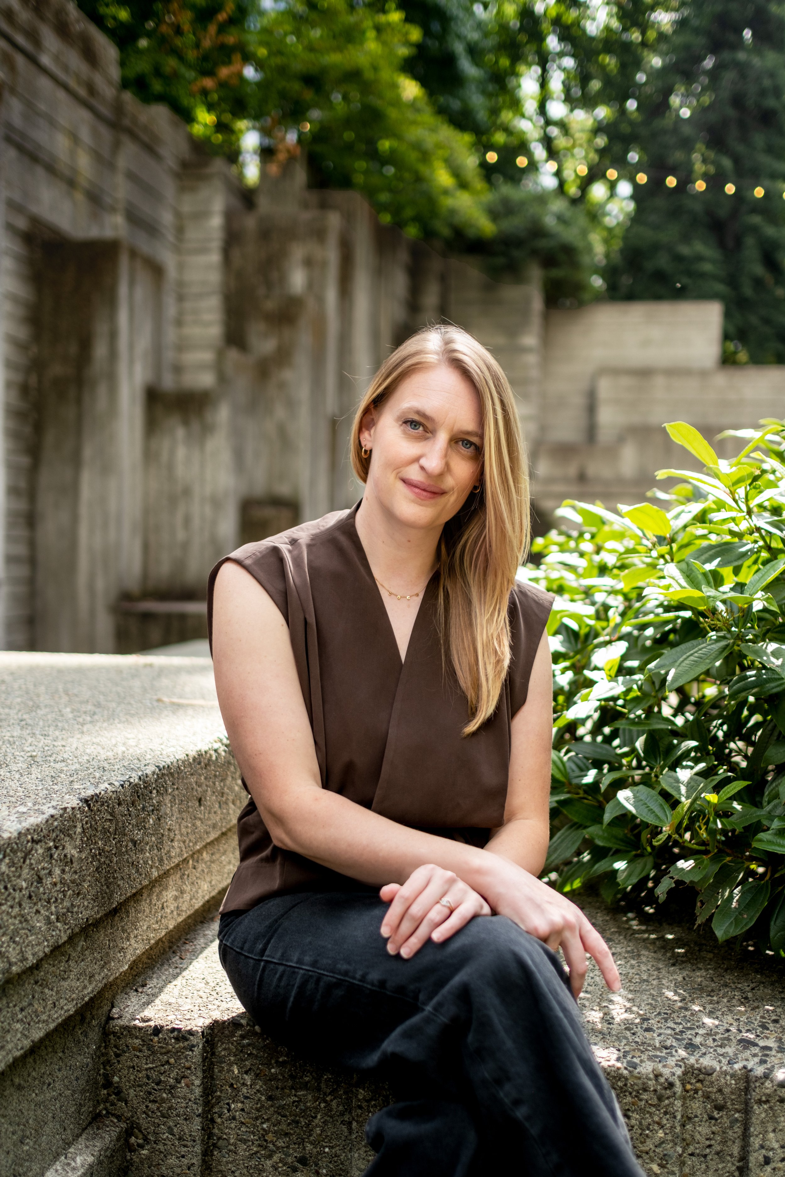 A woman with blonde hair, dressed in a brown top and black pants, sitting on concrete steps outdoors, surrounded by green foliage, with string lights in the background.