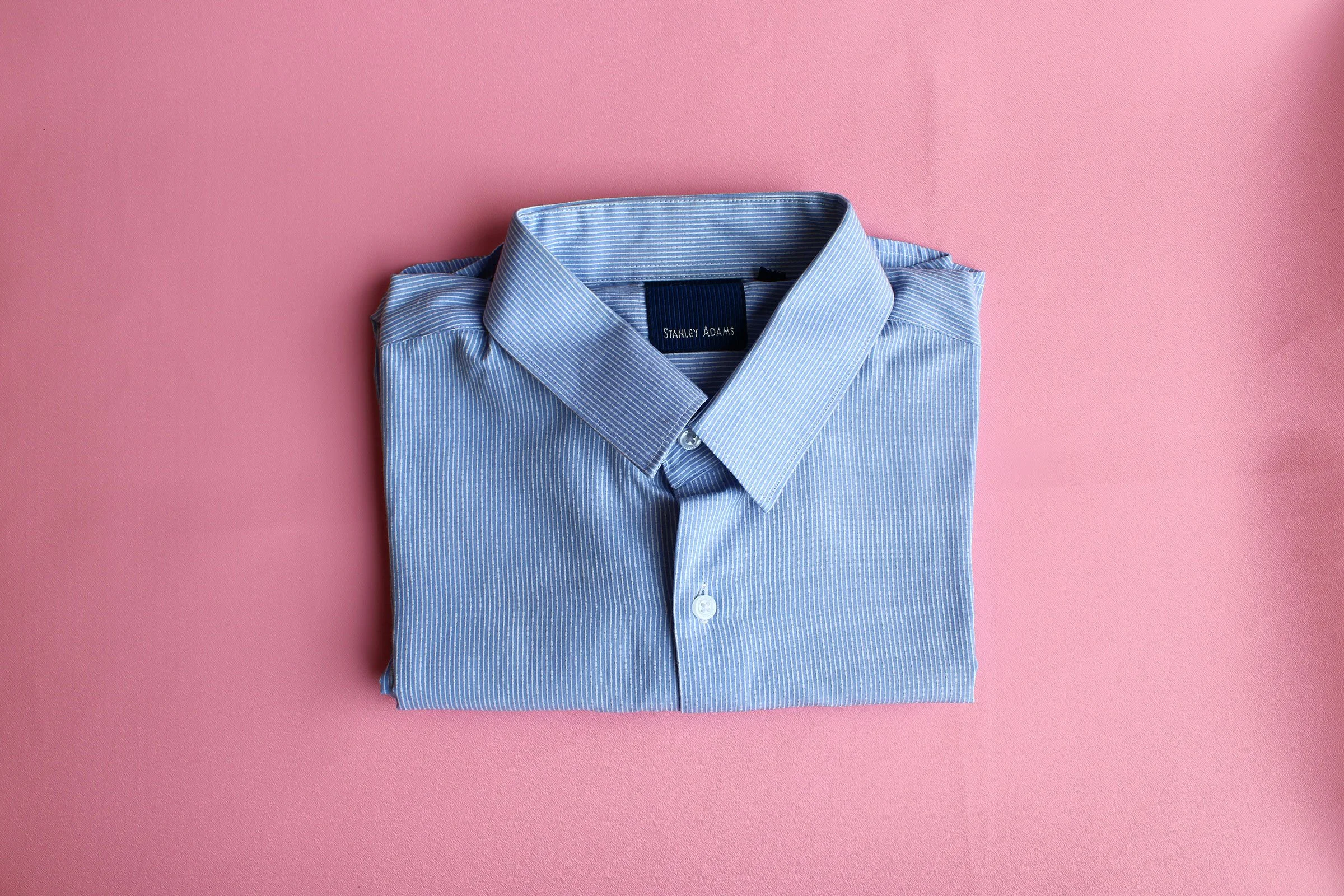 A folded blue button down shirt against a pink background.