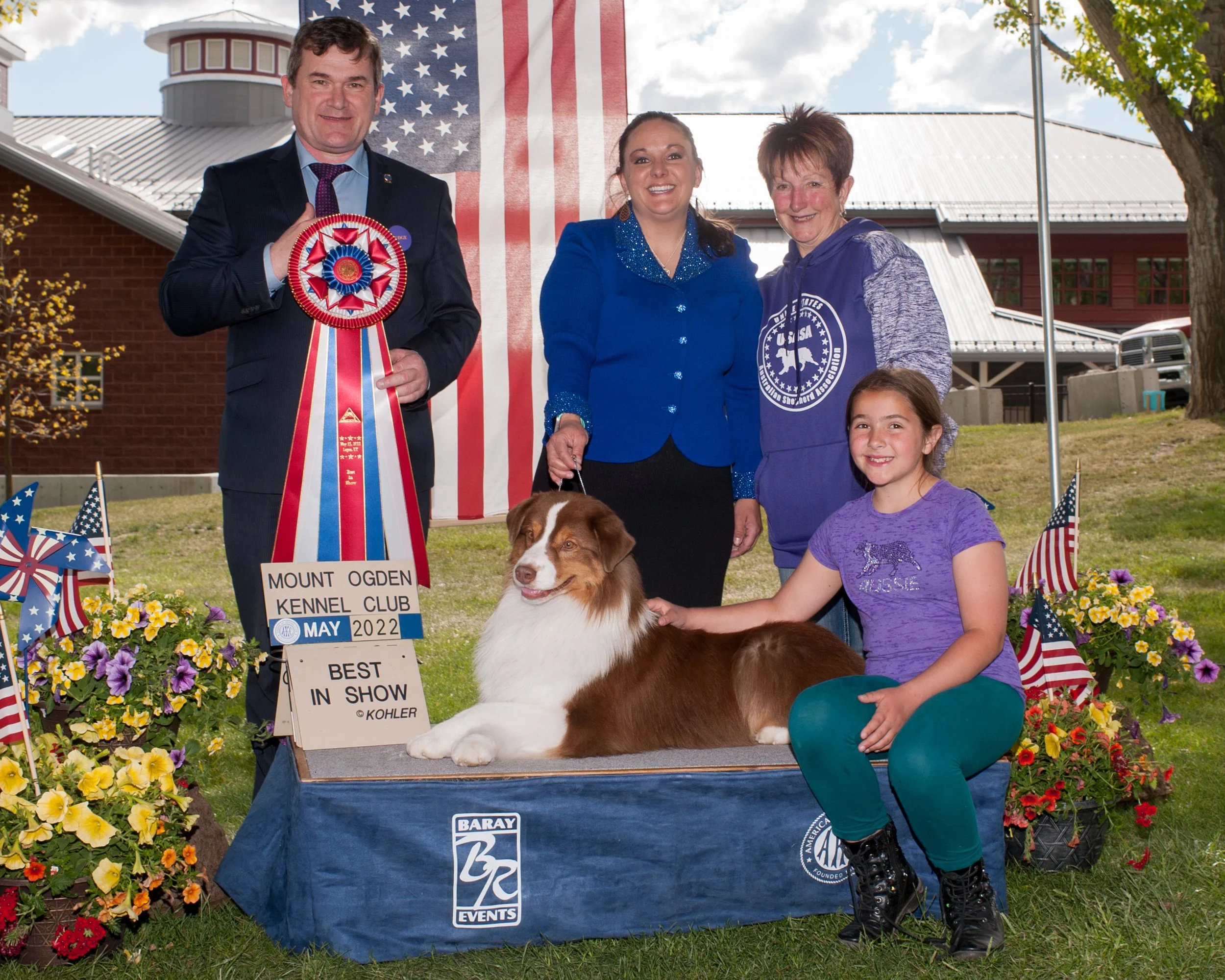 A group of people, including a young girl, posing with a dog that has won 'Best in Show' at the Mount Ogden Kennel Club in May 2022. The man on the left holds a large ribbon; the girl and two women are smiling, with the dog lying on a platform. The b