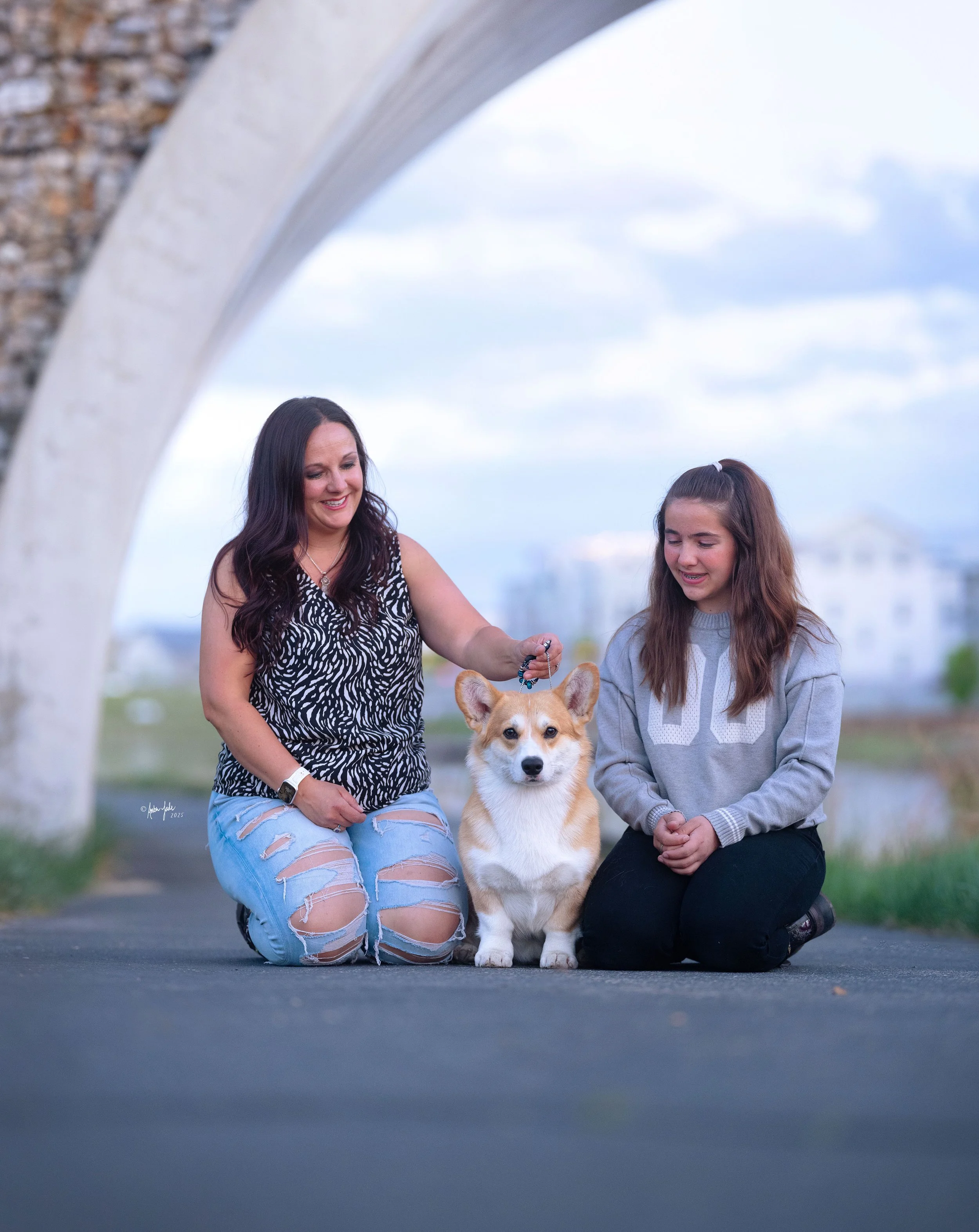 A woman and a girl kneeling on a paved path with a Corgi at Daybreak Lake in Utah. They are under a stone bridge, with water and buildings in the background.
