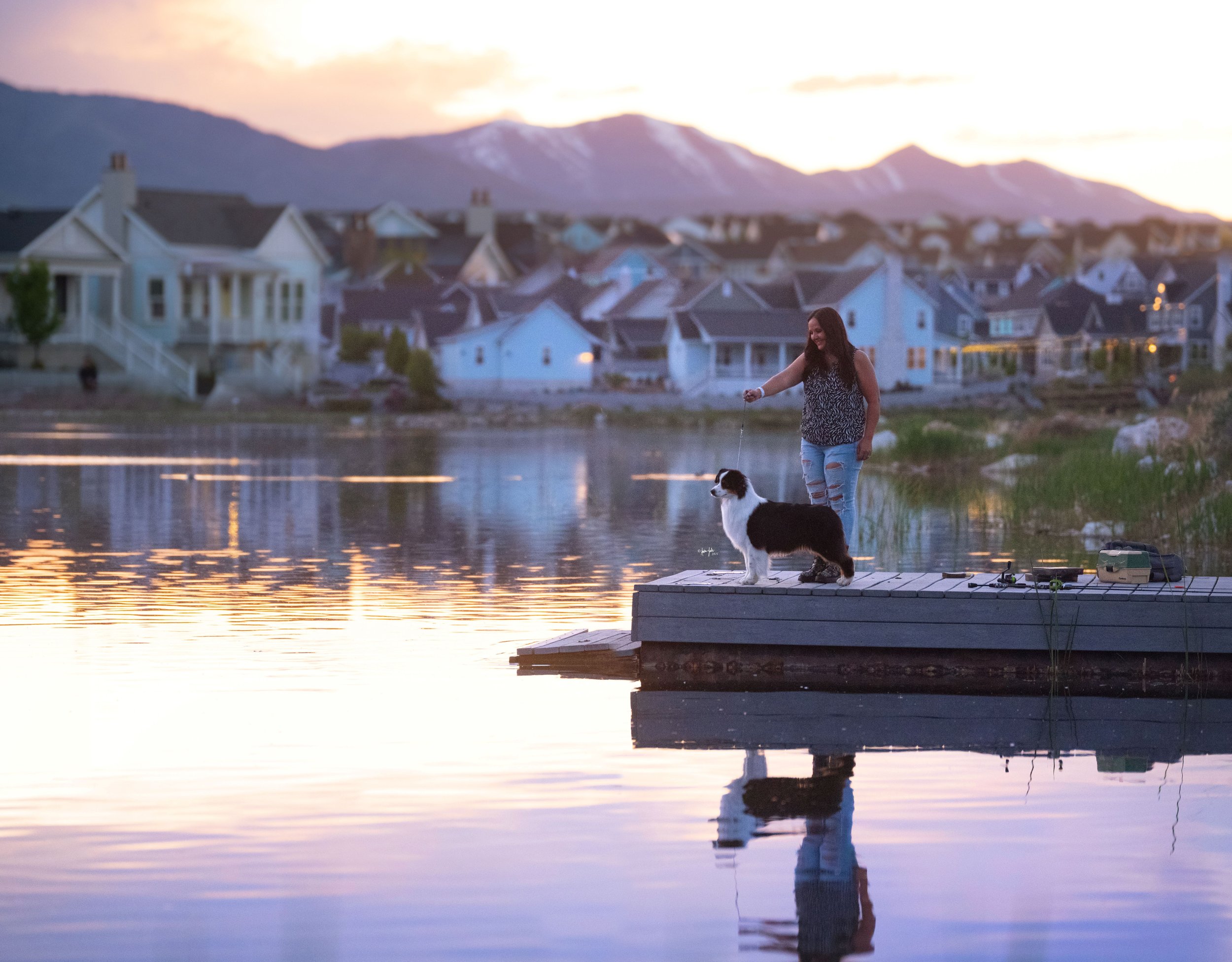 A woman and her black tri Australian Shepherd dog stand on a dock at Daybreak Lake next to a calm lake at sunset, with a backdrop of houses and mountains.