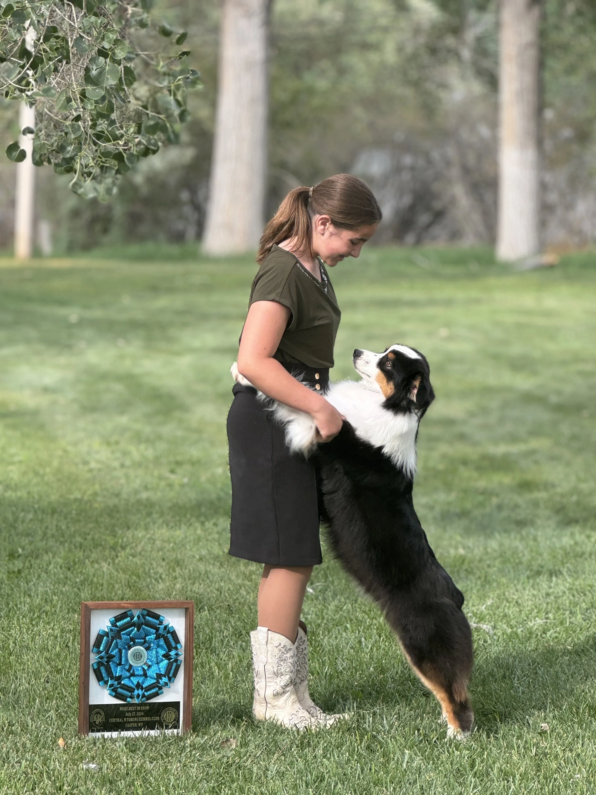 Young woman in black skirt and white cowboy boots standing on grass hugging her black tri Australian Shepherd, on its hind legs looking at her. There is an award ribbon and plaque on the ground to the left.