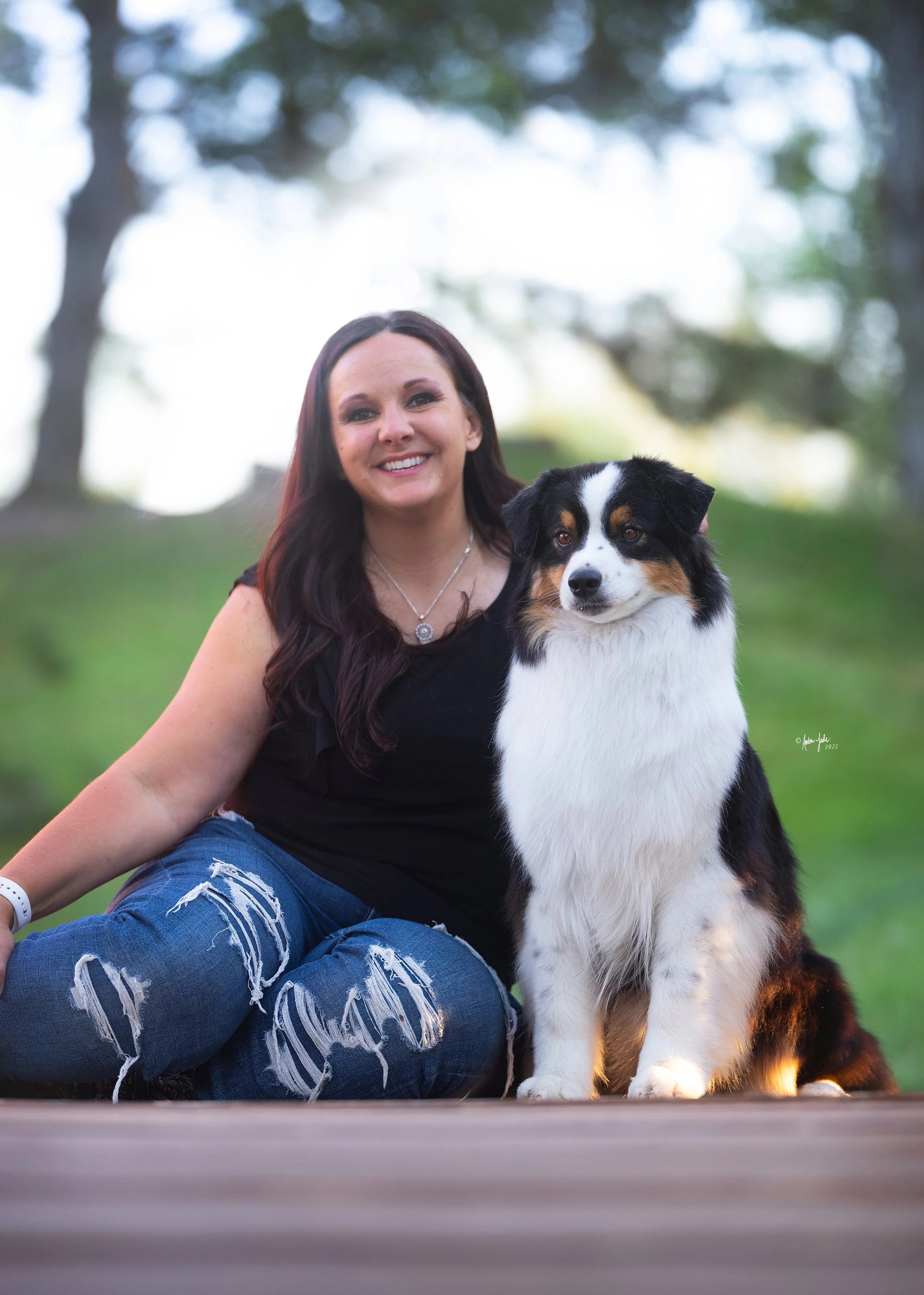 A woman with long dark hair, wearing a black sleeveless top and ripped jeans, sitting outdoors on a wooden surface at International Peace Gardens next to an Australian Shepherd dog with a tri color coat, in a park with trees in the background.